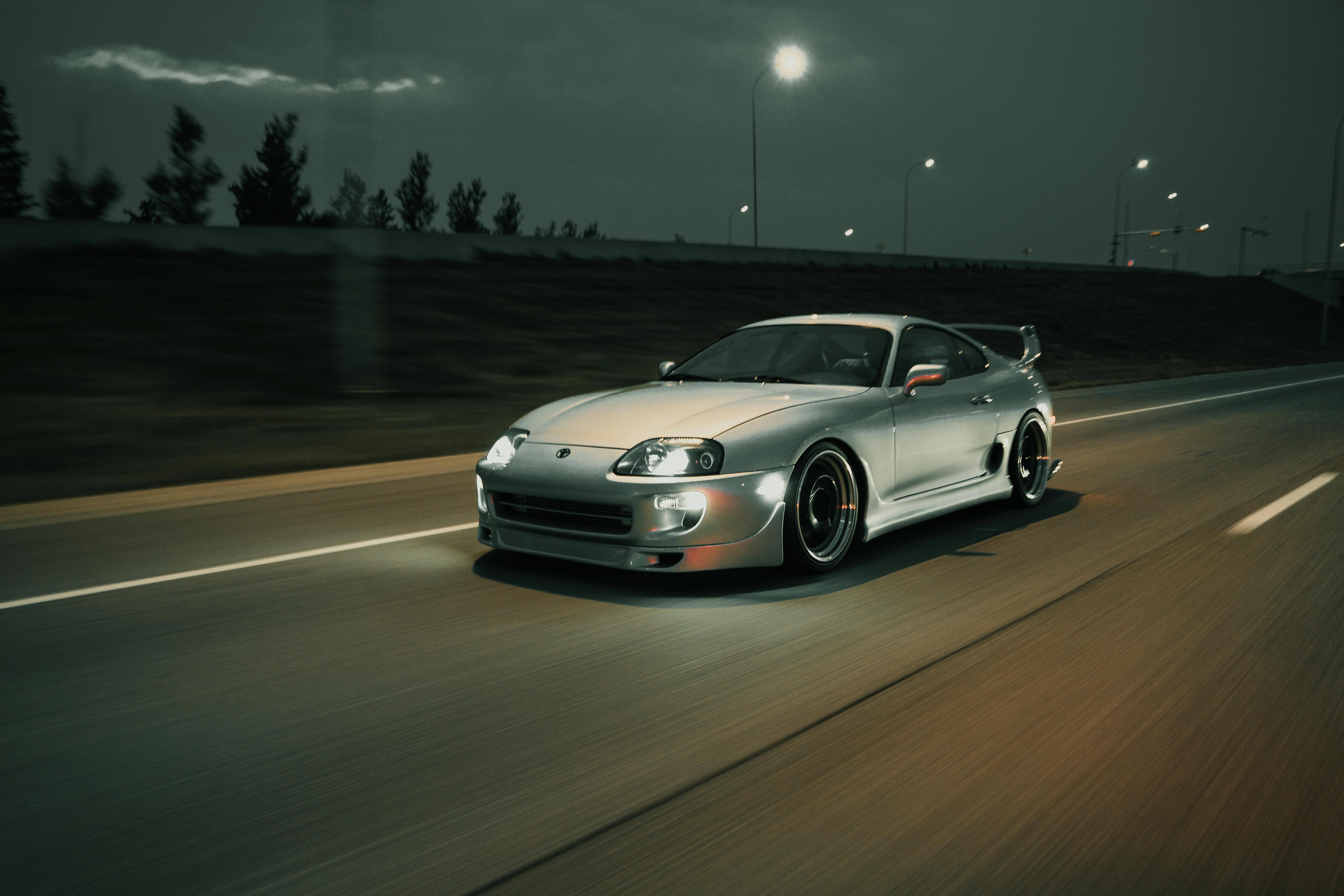 A motion-blur shot of a silver Toyota Supra driving at night on a dark highway, illuminated by streetlights.
