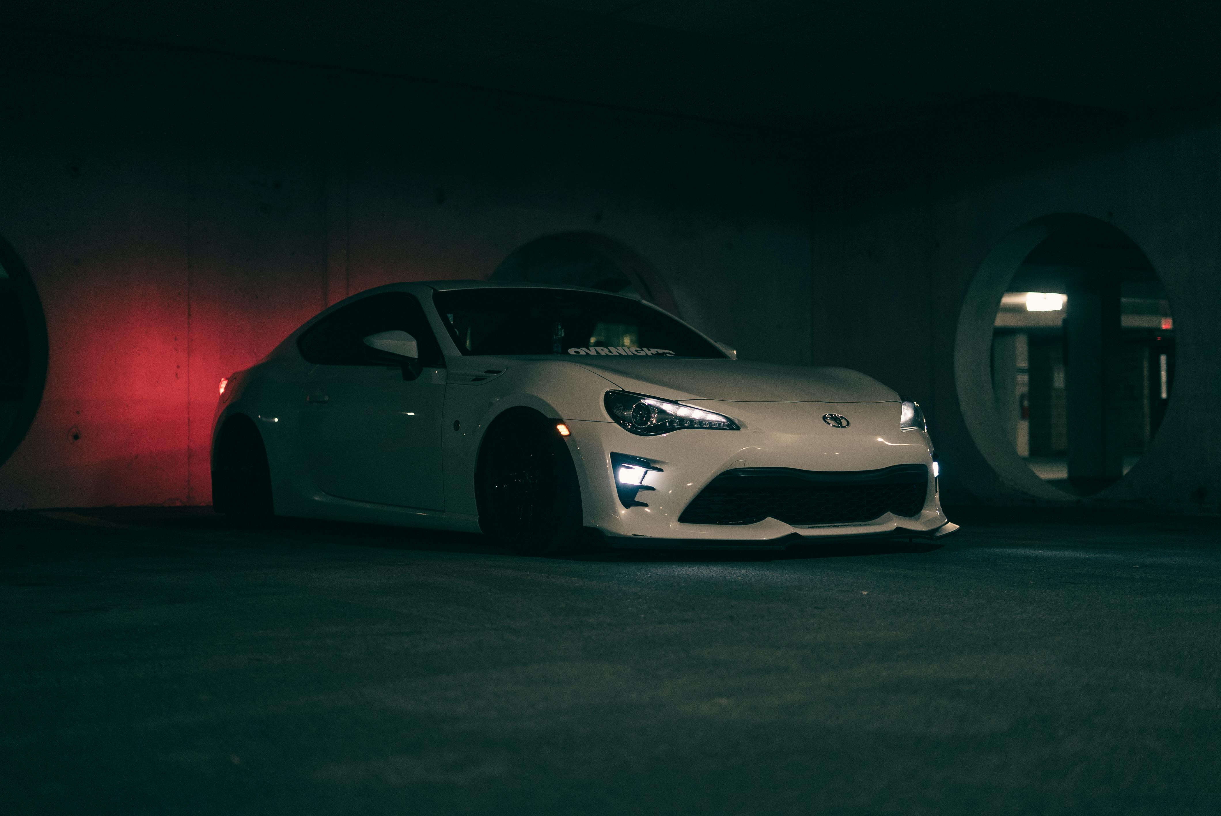 A white Toyota GR86 parked in a dark, atmospheric concrete structure with a soft red glow in the background.
