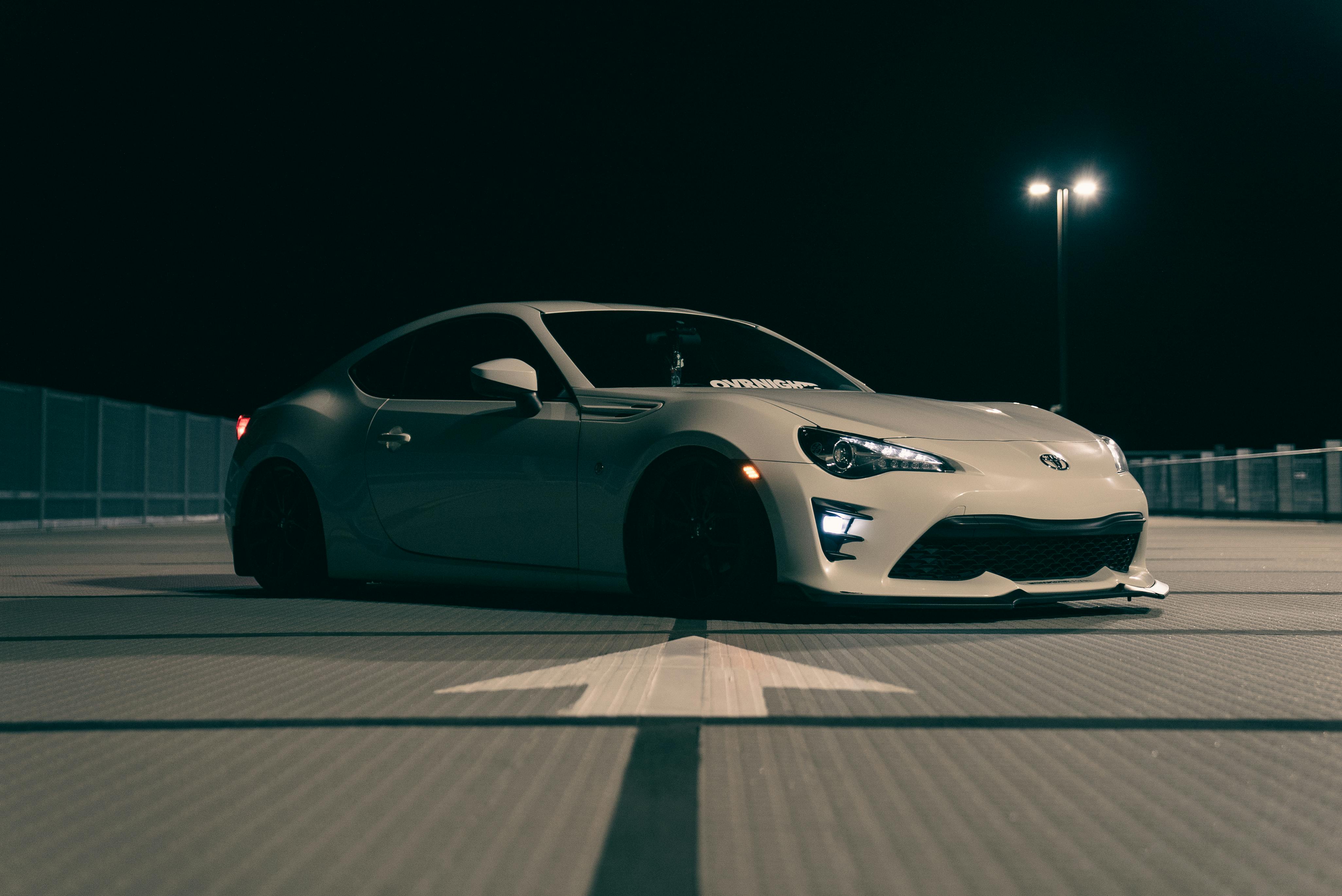 A side profile of a white Toyota GR86 parked on an asphalt surface at night under the bright light of a single street lamp.