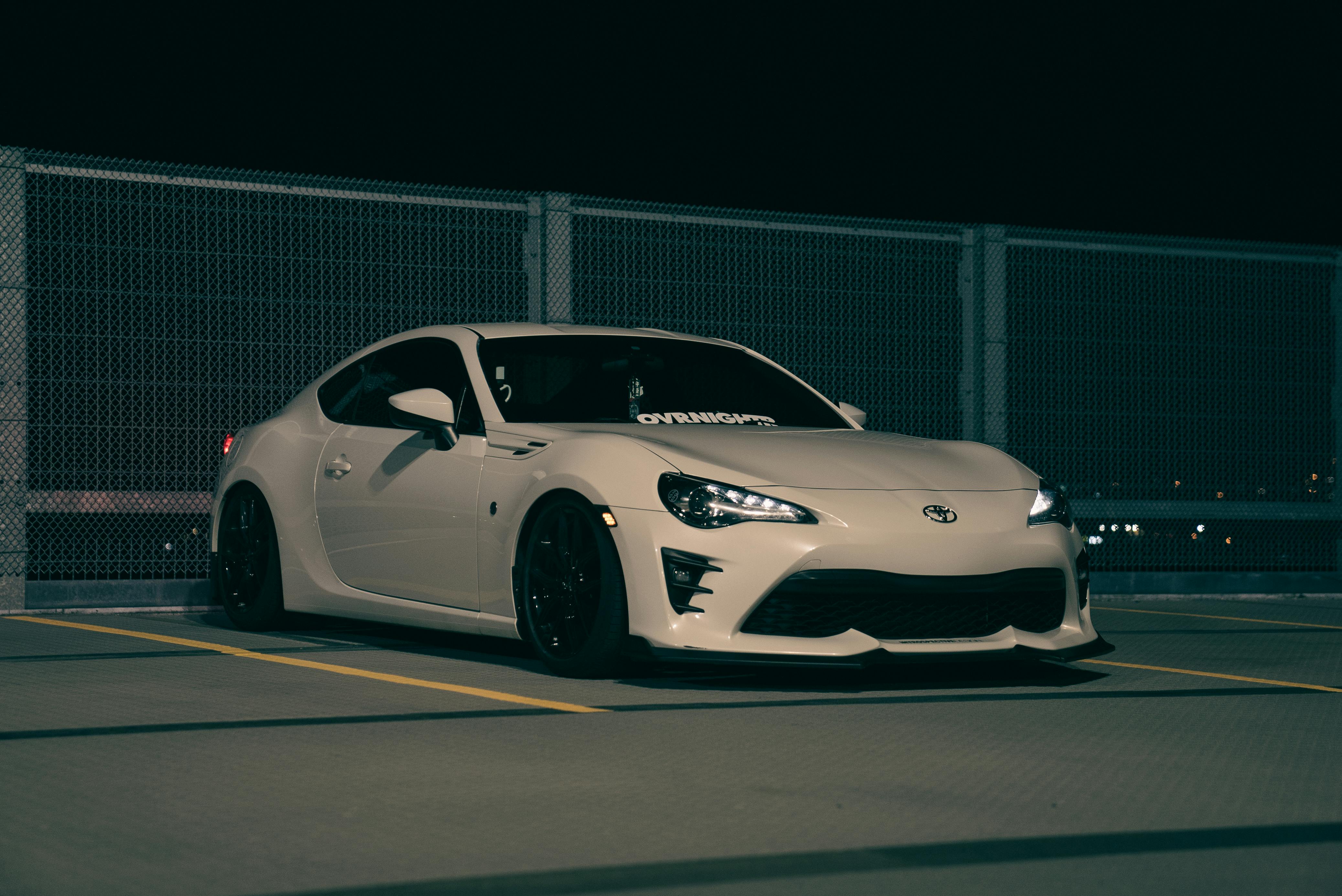 A white Toyota GR86 parked in front of a chain-link fence at night, with a clean, low-profile stance.