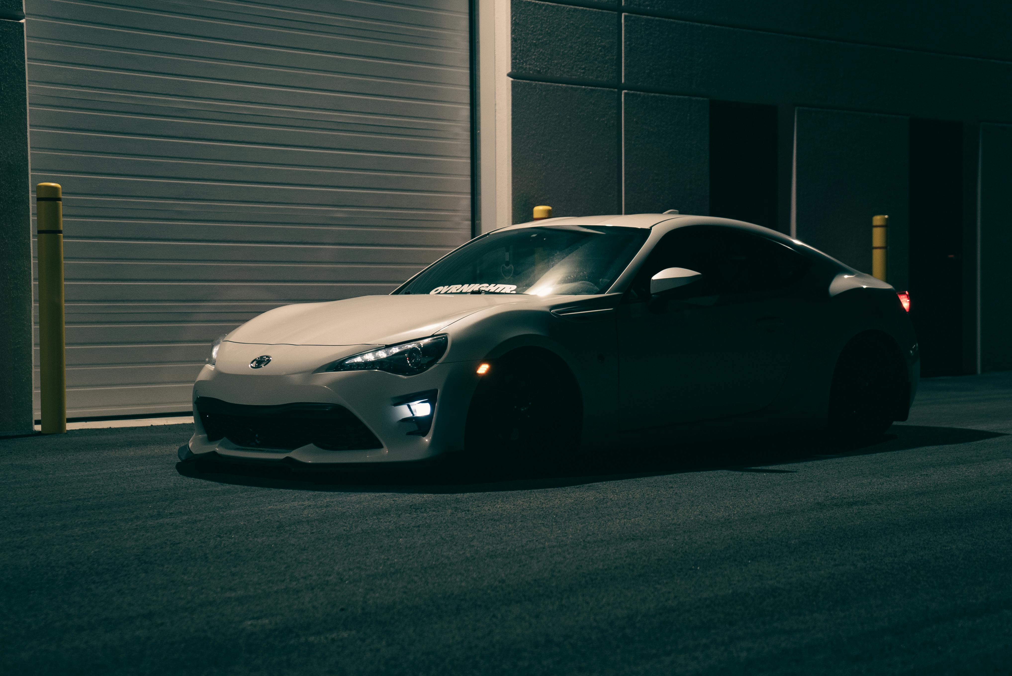 A white Toyota GR86 parked in a dark industrial area next to a closed warehouse garage door.
