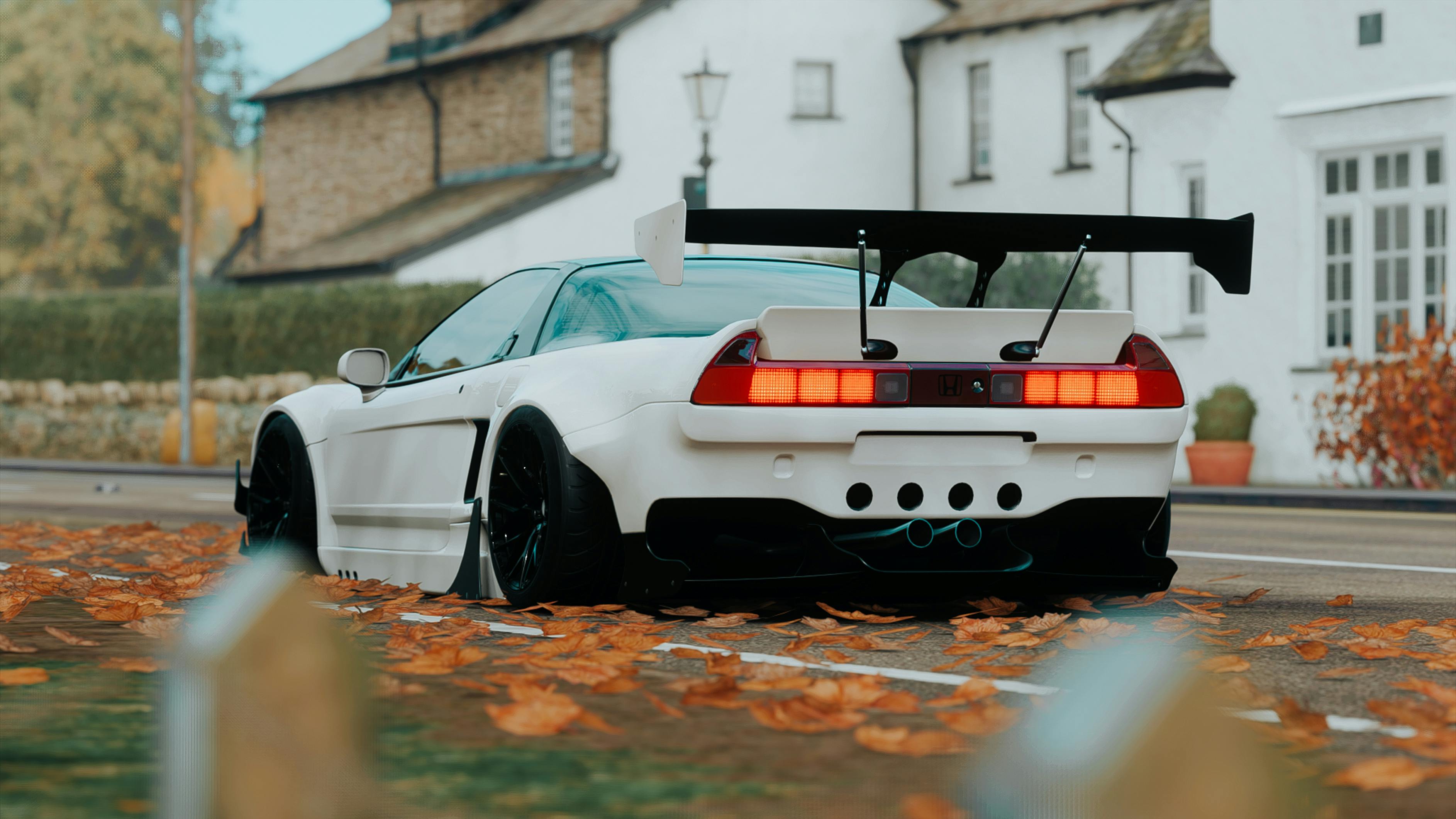 Rear view of a wide-body white Honda NSX with a massive black GT wing, parked on a street covered in orange autumn leaves.