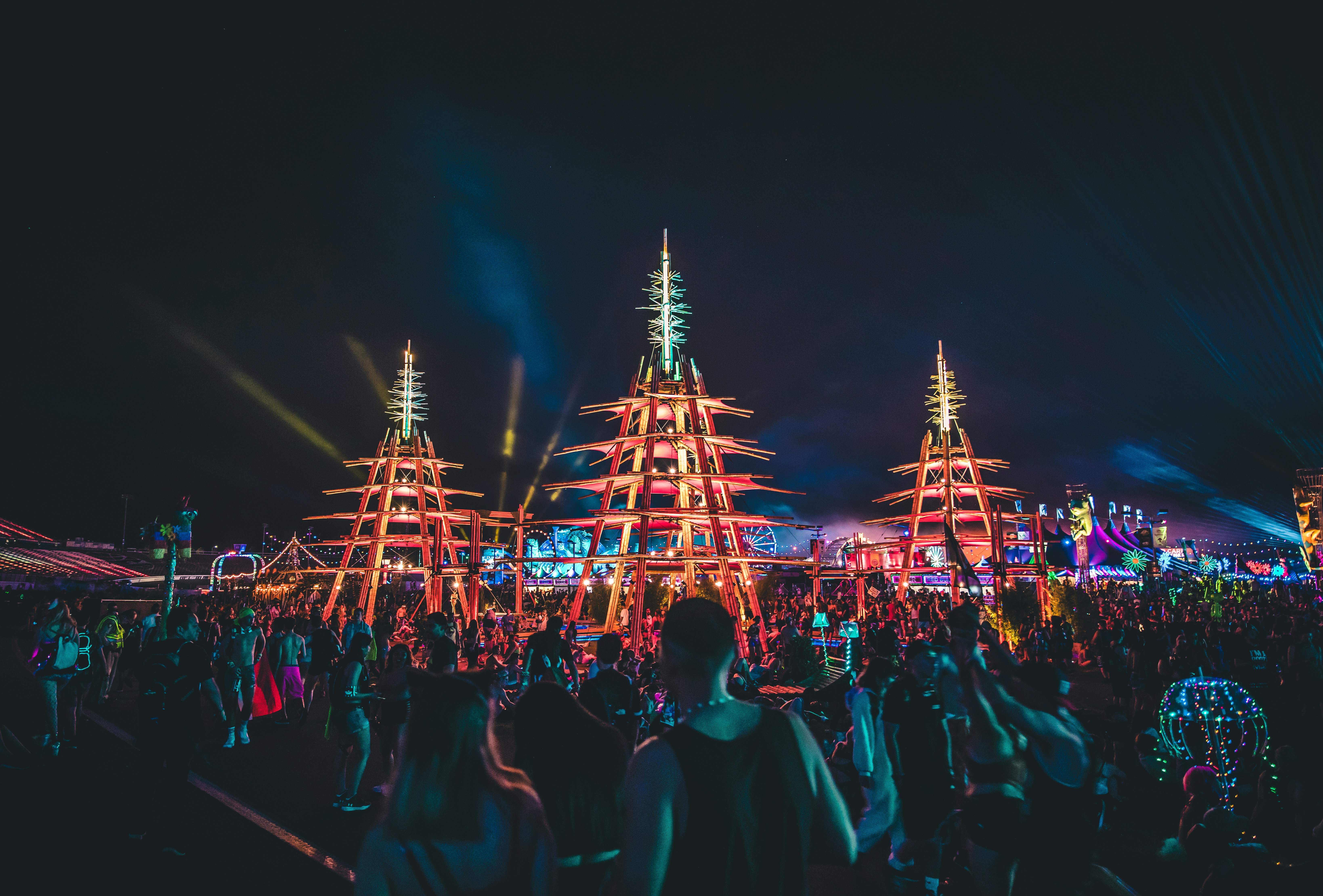 A large outdoor festival at night featuring three towering, illuminated structures shaped like glowing pagodas or trees, surrounded by a massive crowd.