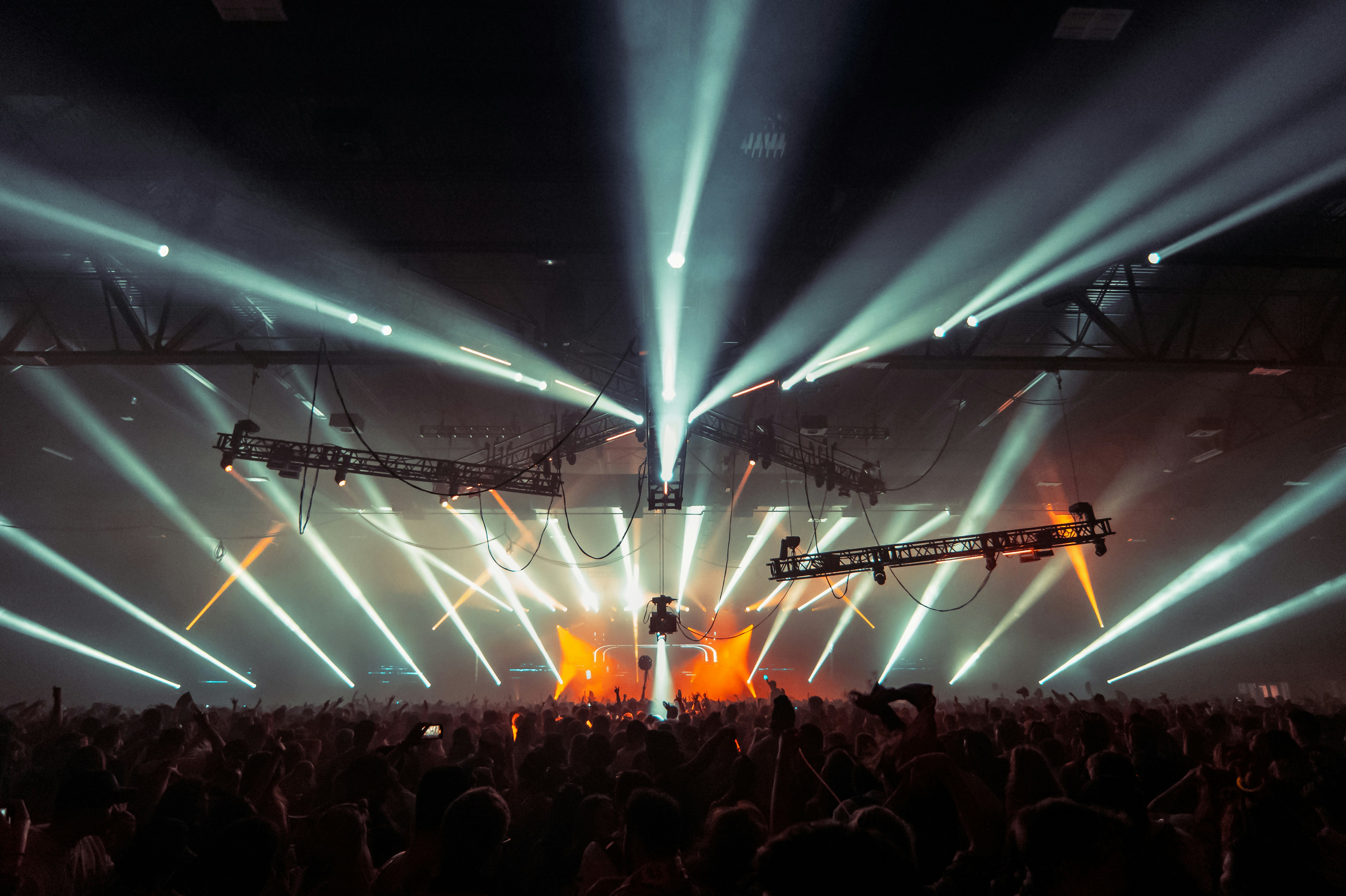 An indoor music concert with a dramatic light show, featuring bright white beams of light radiating from the stage over a dense crowd of people.