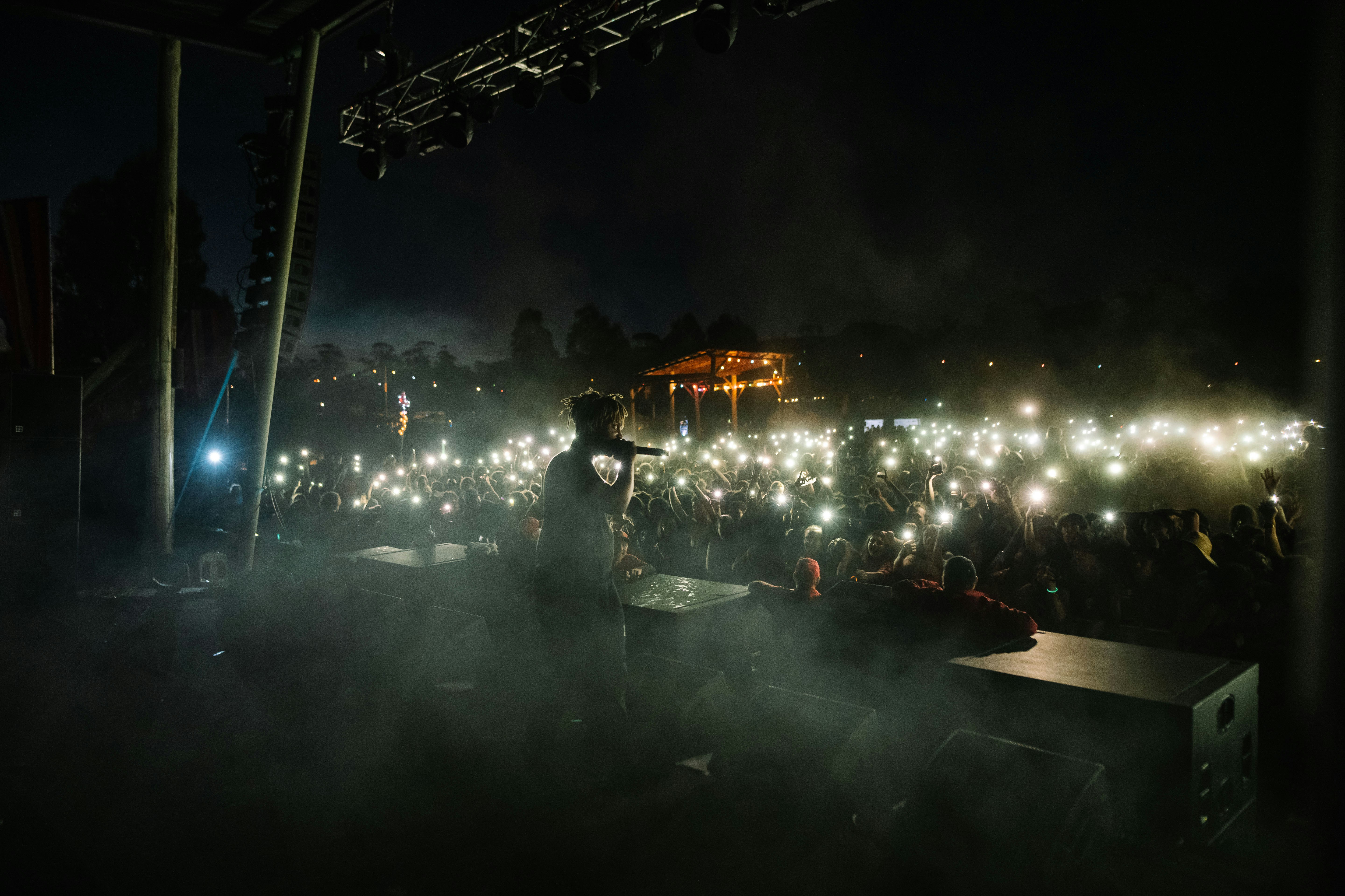 A silhouette of the artist Juice WRLD performing on an outdoor stage at night, looking out at a vast field of glowing lights from the audience's smartphones during a live concert.
