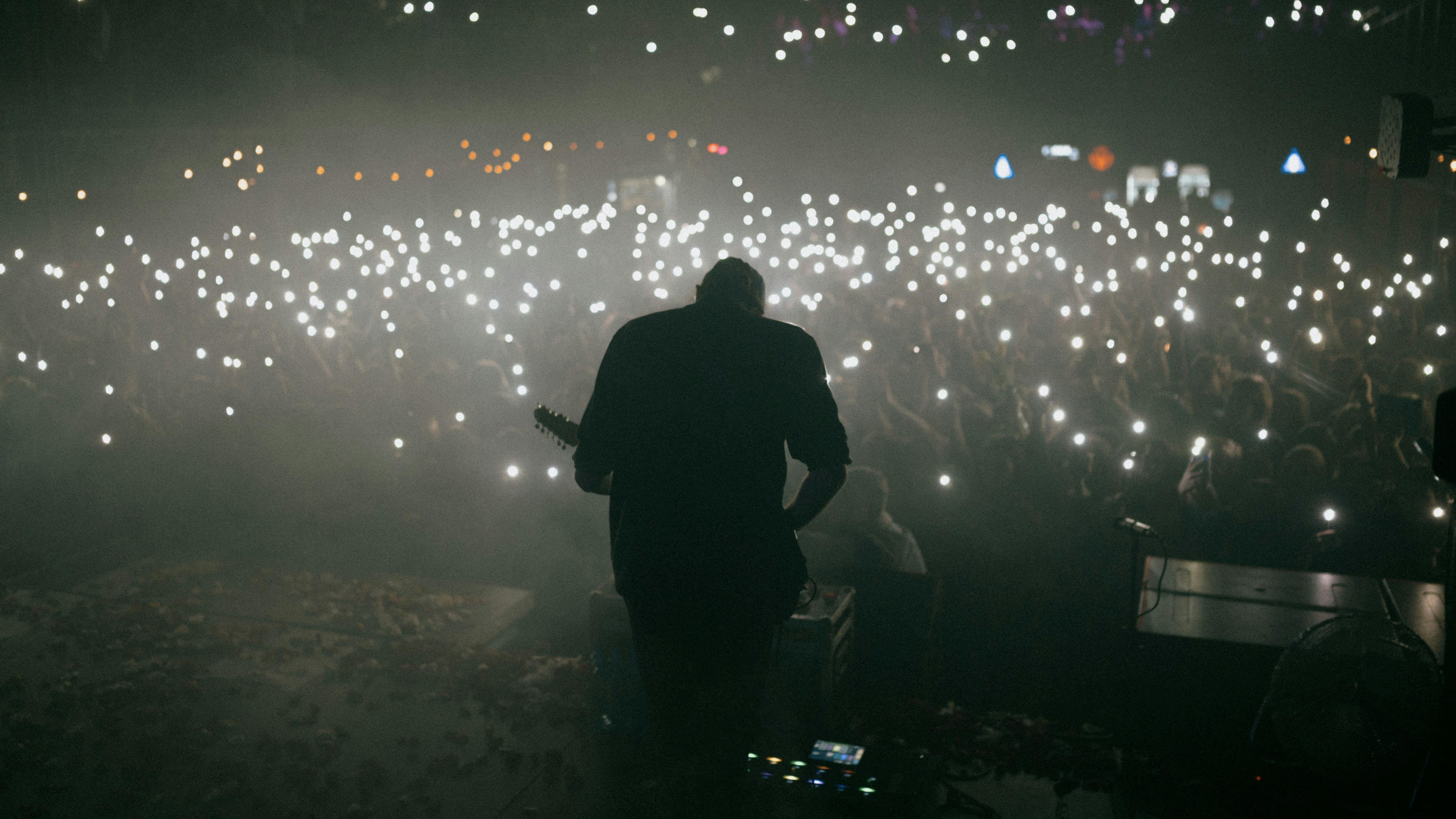 A silhouette of a musician with a guitar on stage, facing a vast audience that has illuminated the dark venue with hundreds of phone flashlights.