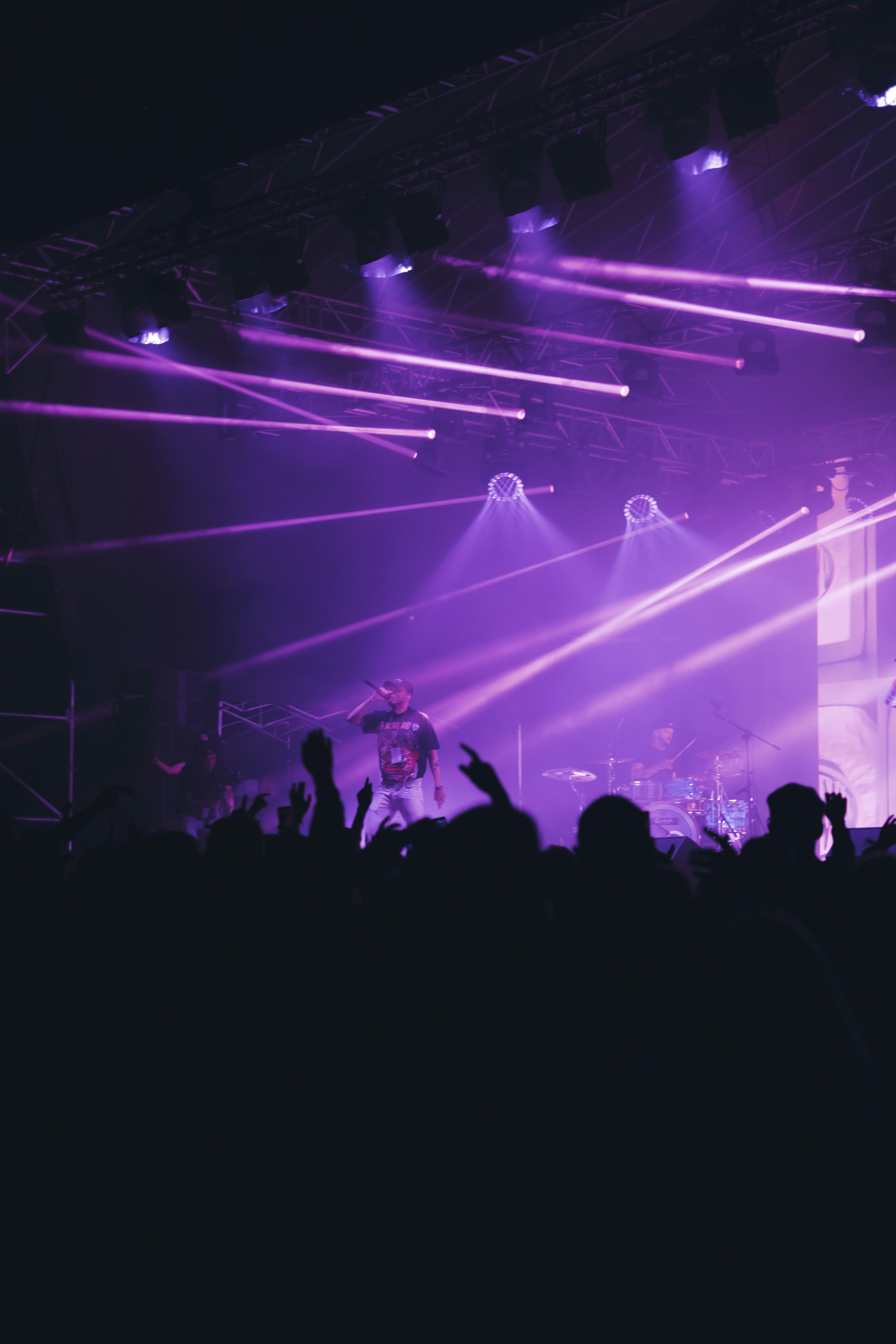 A performer on stage at a concert, silhouetted against vibrant purple laser lights and hazy smoke as the crowd raises their hands.
