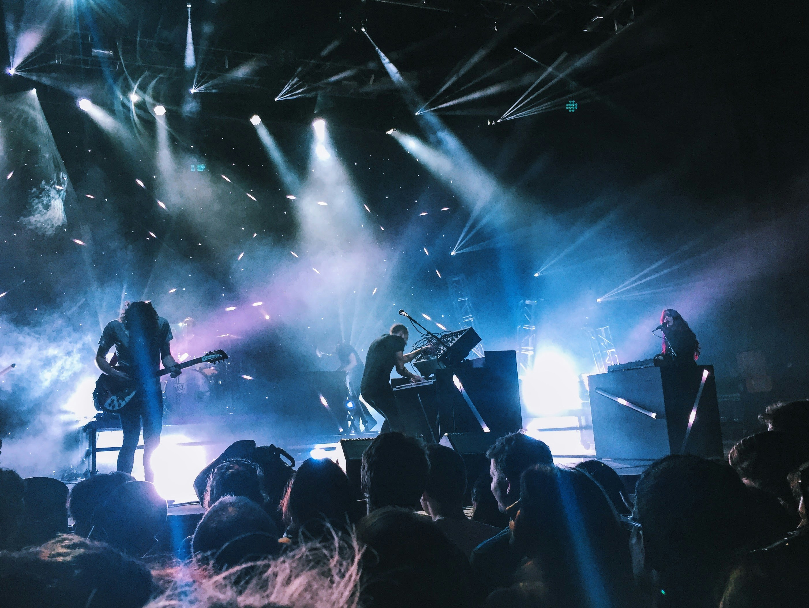 A high-energy rock band performing on stage under intense blue and white spotlights, with silhouettes of the guitarist and keyboardist in action.