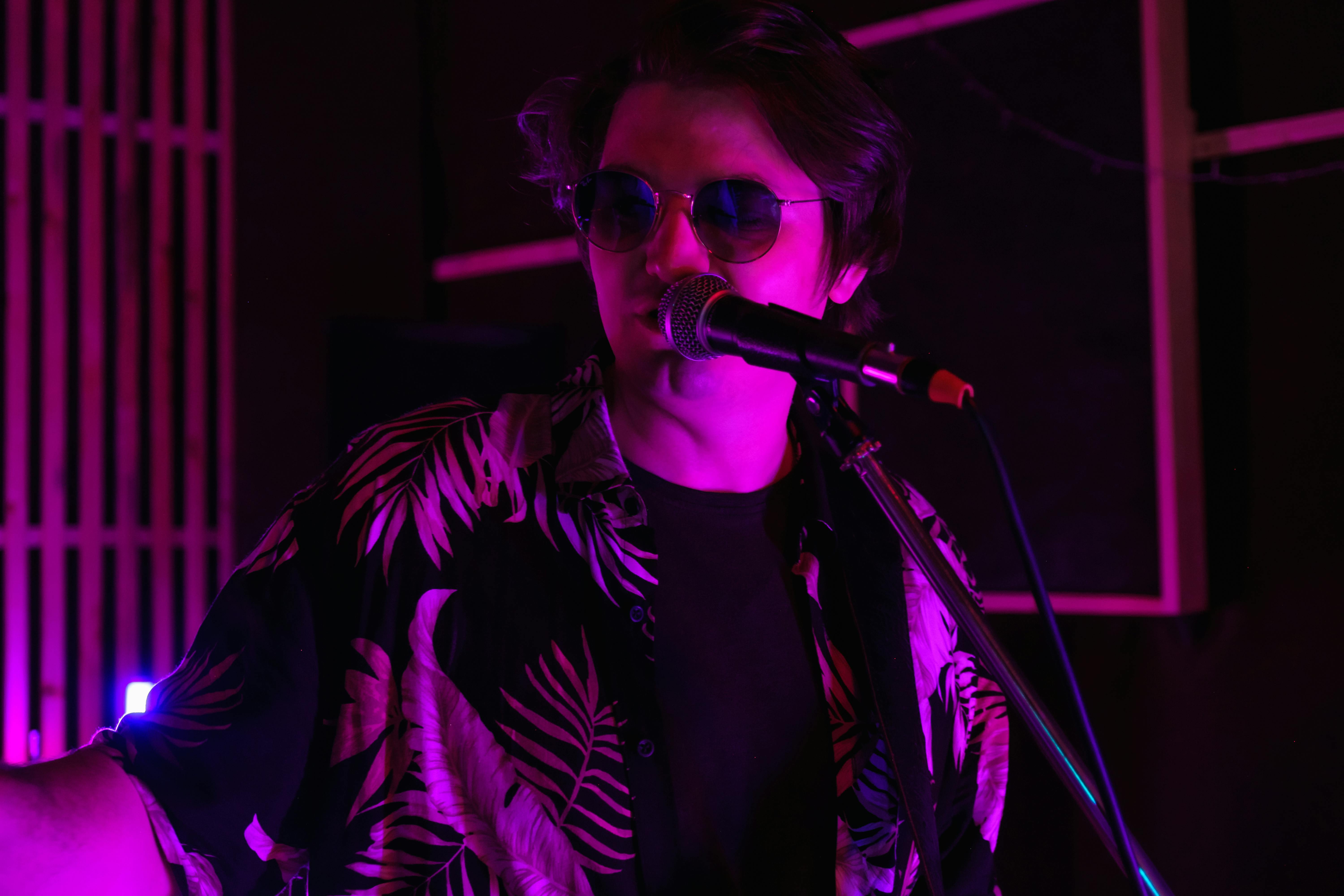 A male performer with sunglasses and a patterned black shirt singing into a microphone under vibrant purple and pink stage lights.