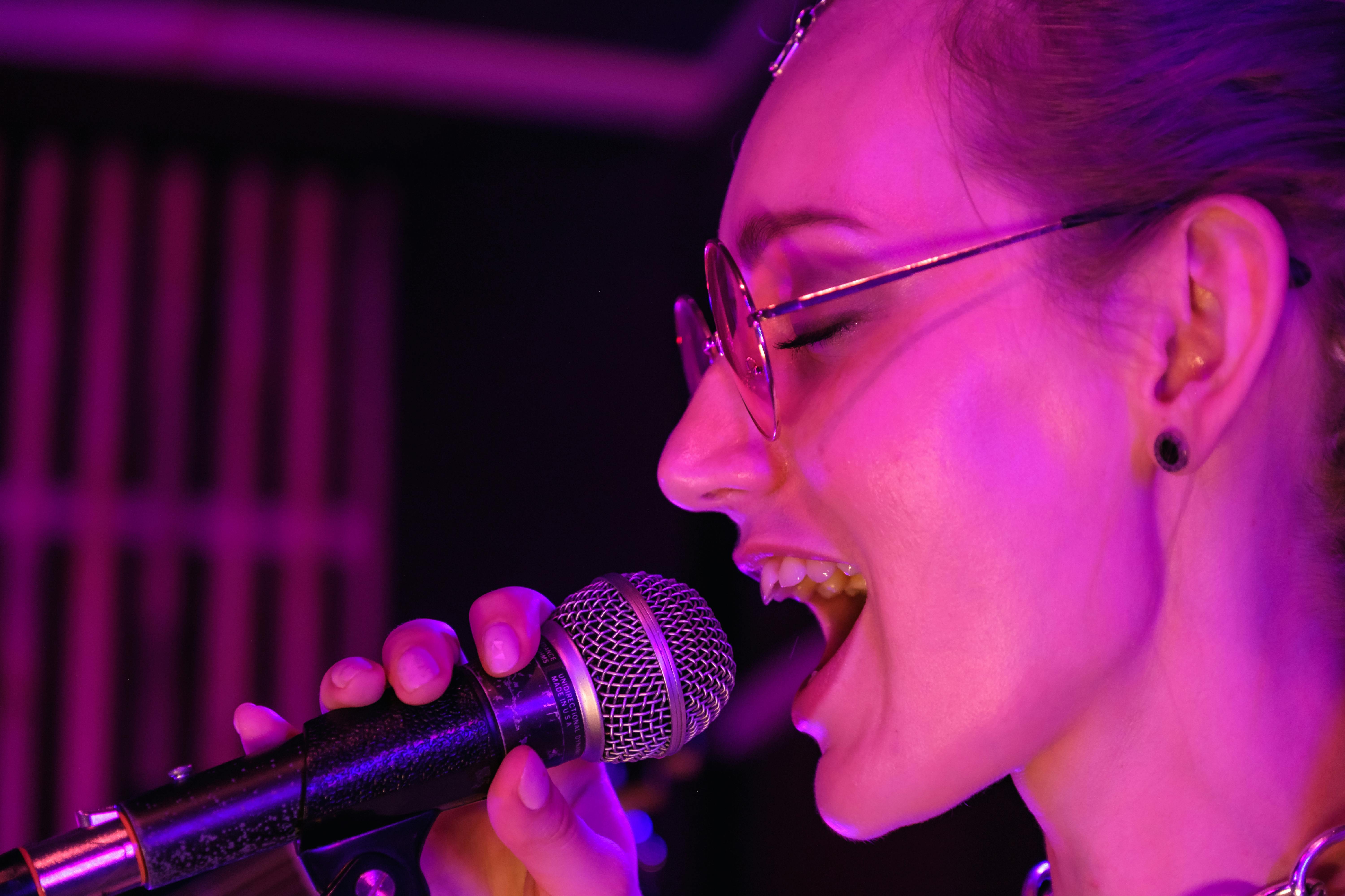 A close-up profile of a female singer with glasses performing passionately into a microphone, bathed in bright pink neon light.