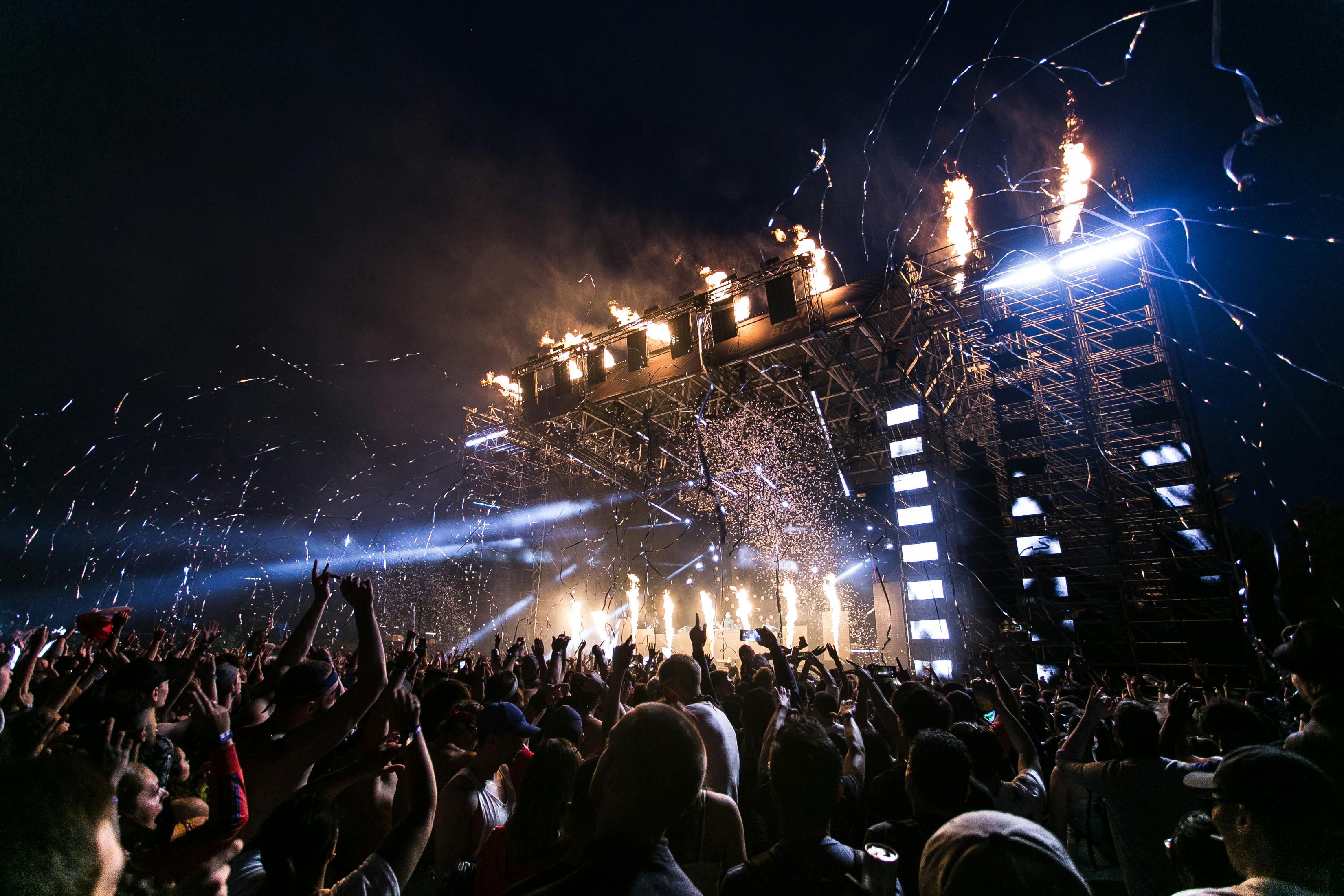 A massive outdoor concert stage at night featuring intense pyrotechnics and sparks flying over a large, cheering crowd.