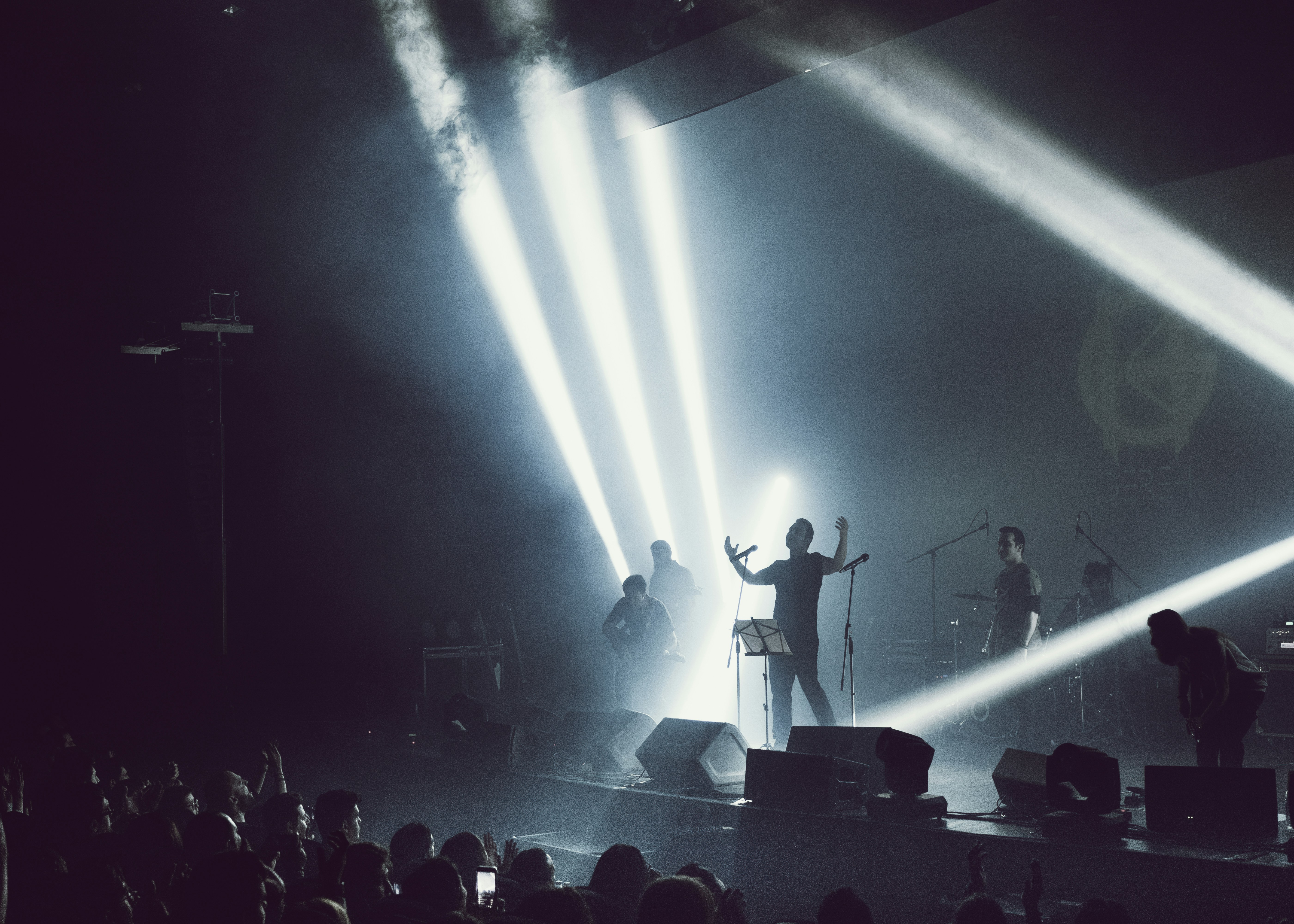 A band performing on stage silhouetted against dramatic, wide beams of white spotlight and hazy stage smoke.