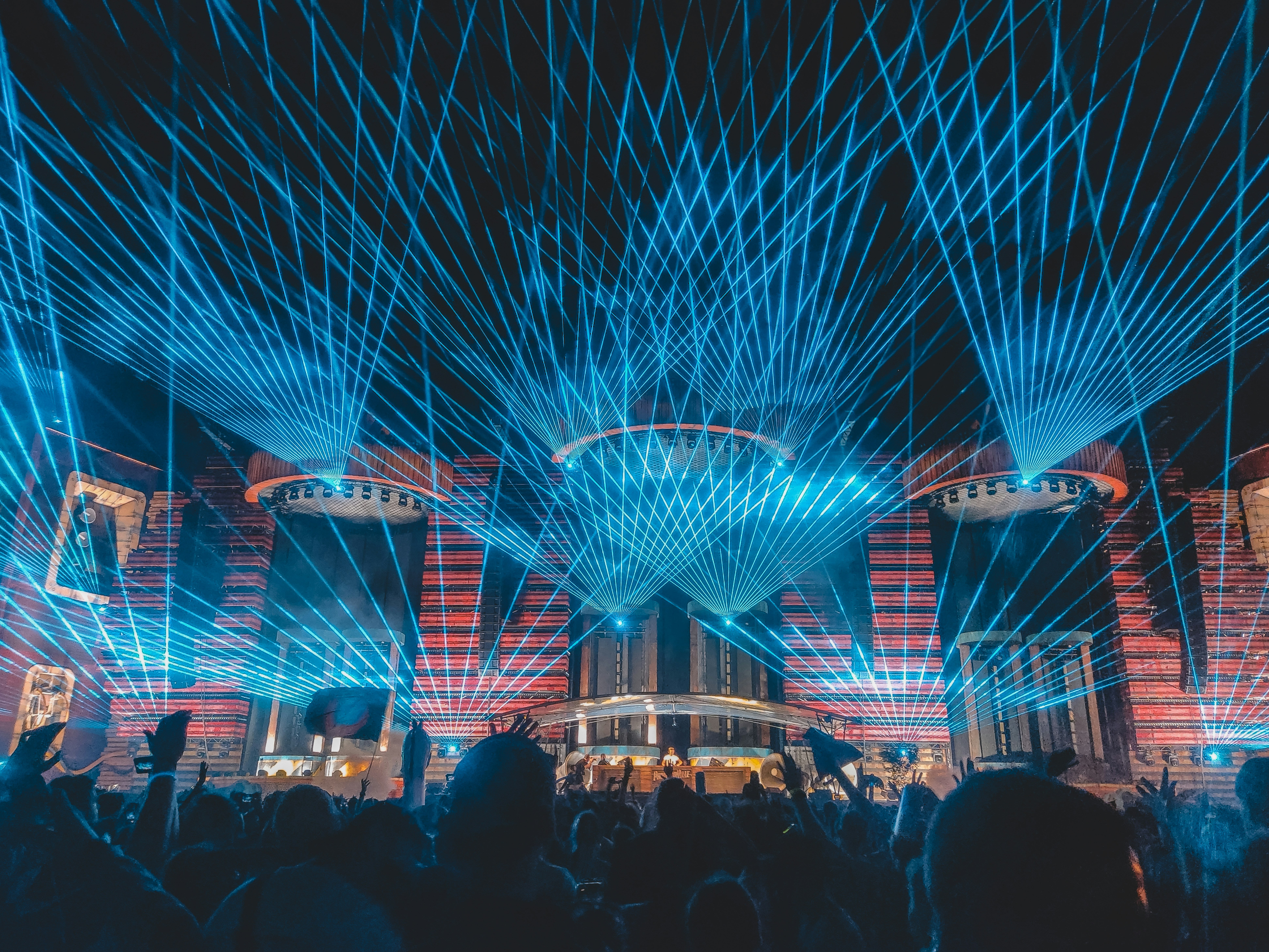 A high-energy electronic music event with a complex web of bright blue laser lights crisscrossing over a dense audience.