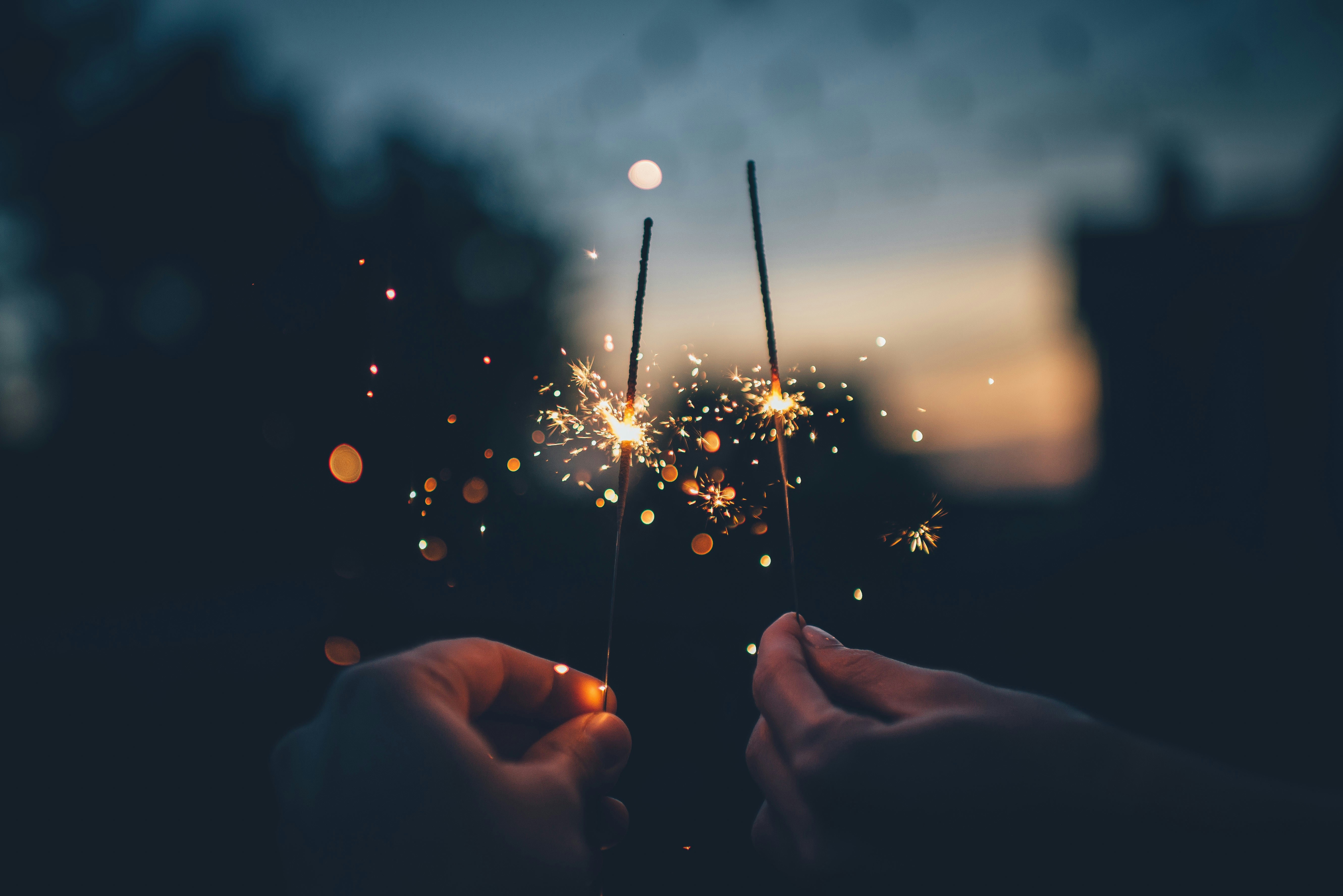 Close up of hands holding two glowing, sparking sparklers against a soft, blurred sunset background.