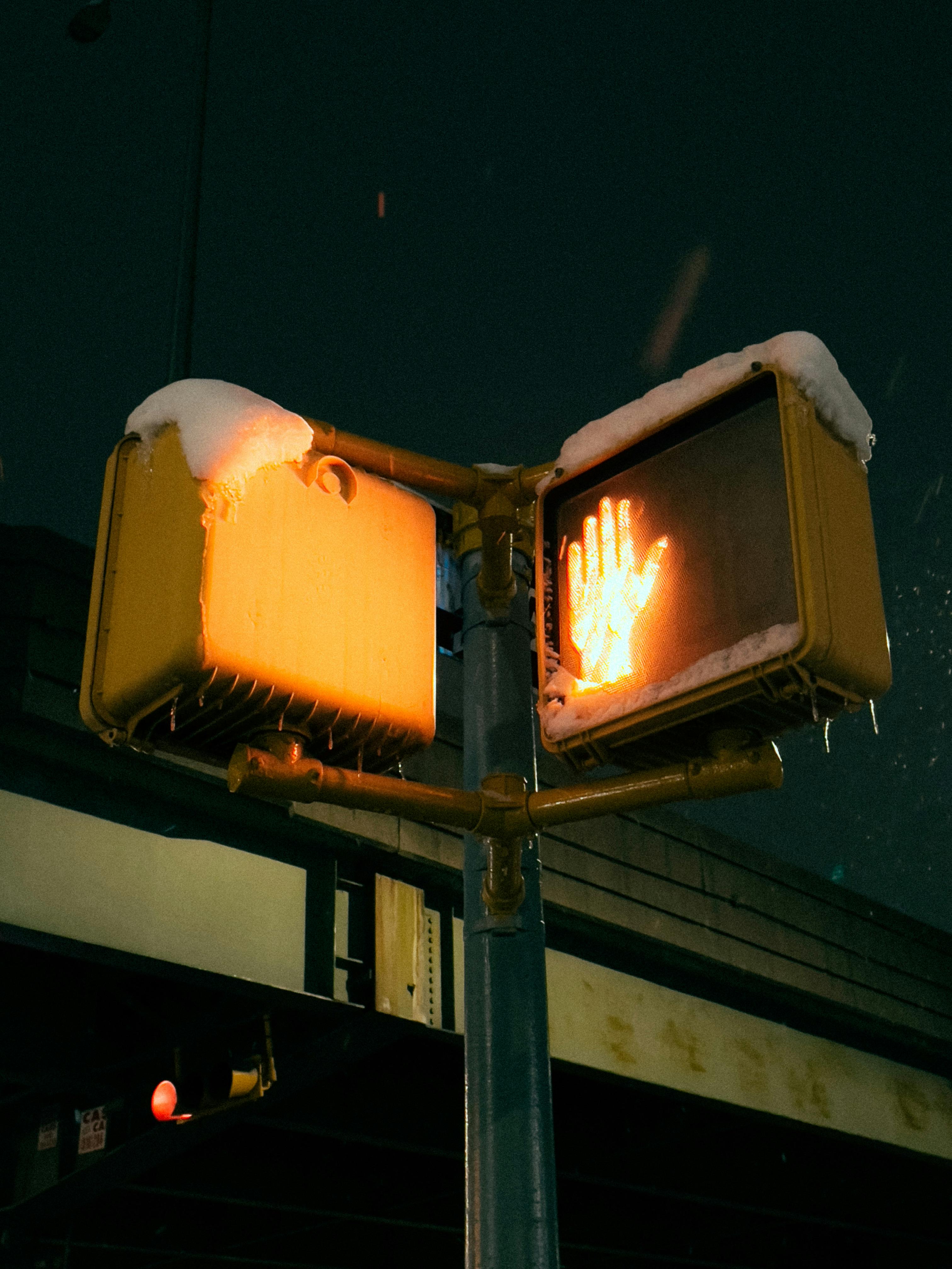 Snow covered pedestrian signal at night, showing a glowing orange "Don't Walk" hand icon against a dark, moody street background.