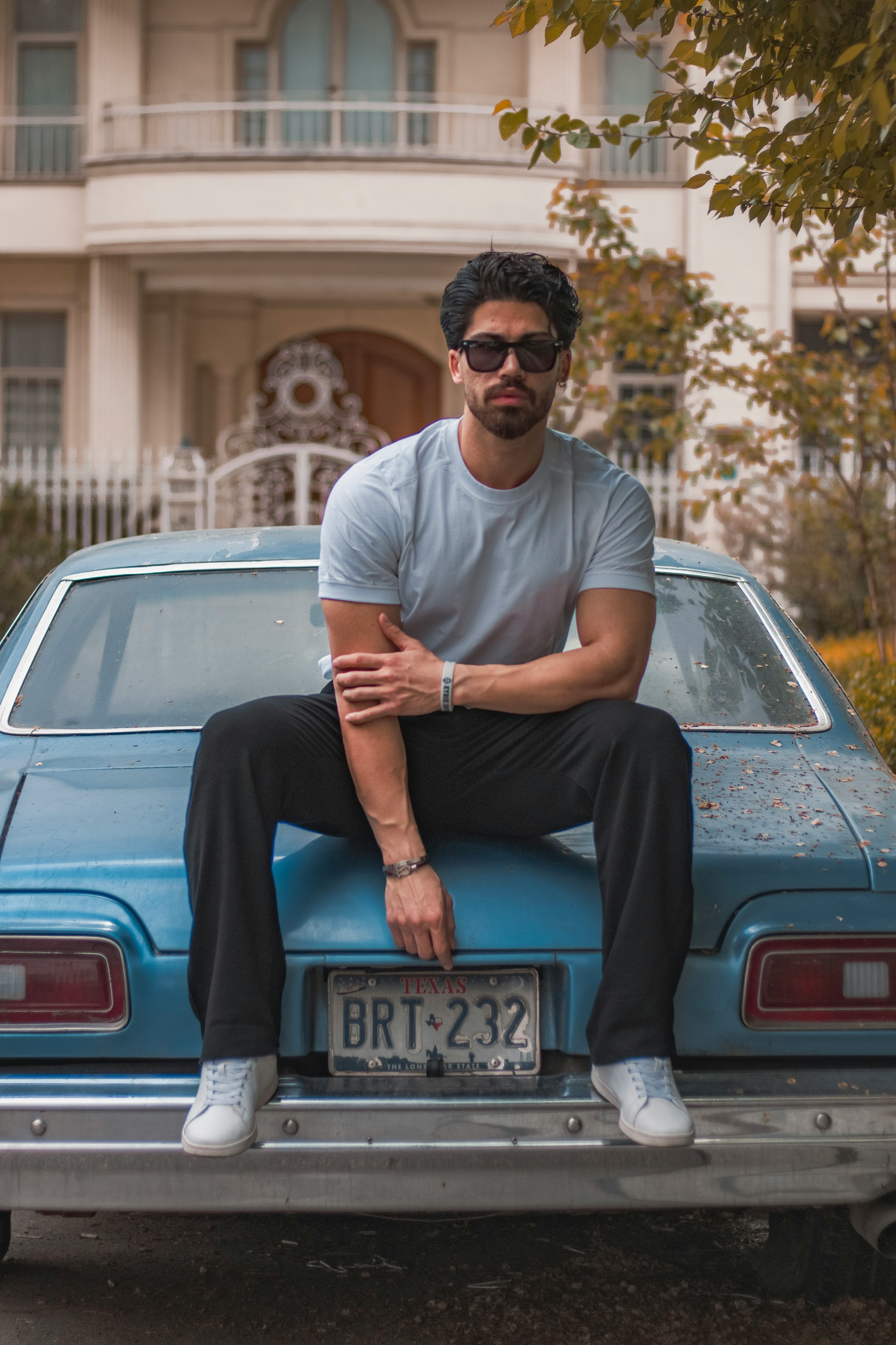A man wearing a grey t-shirt and sunglasses sitting casually on the trunk of a vintage blue car.