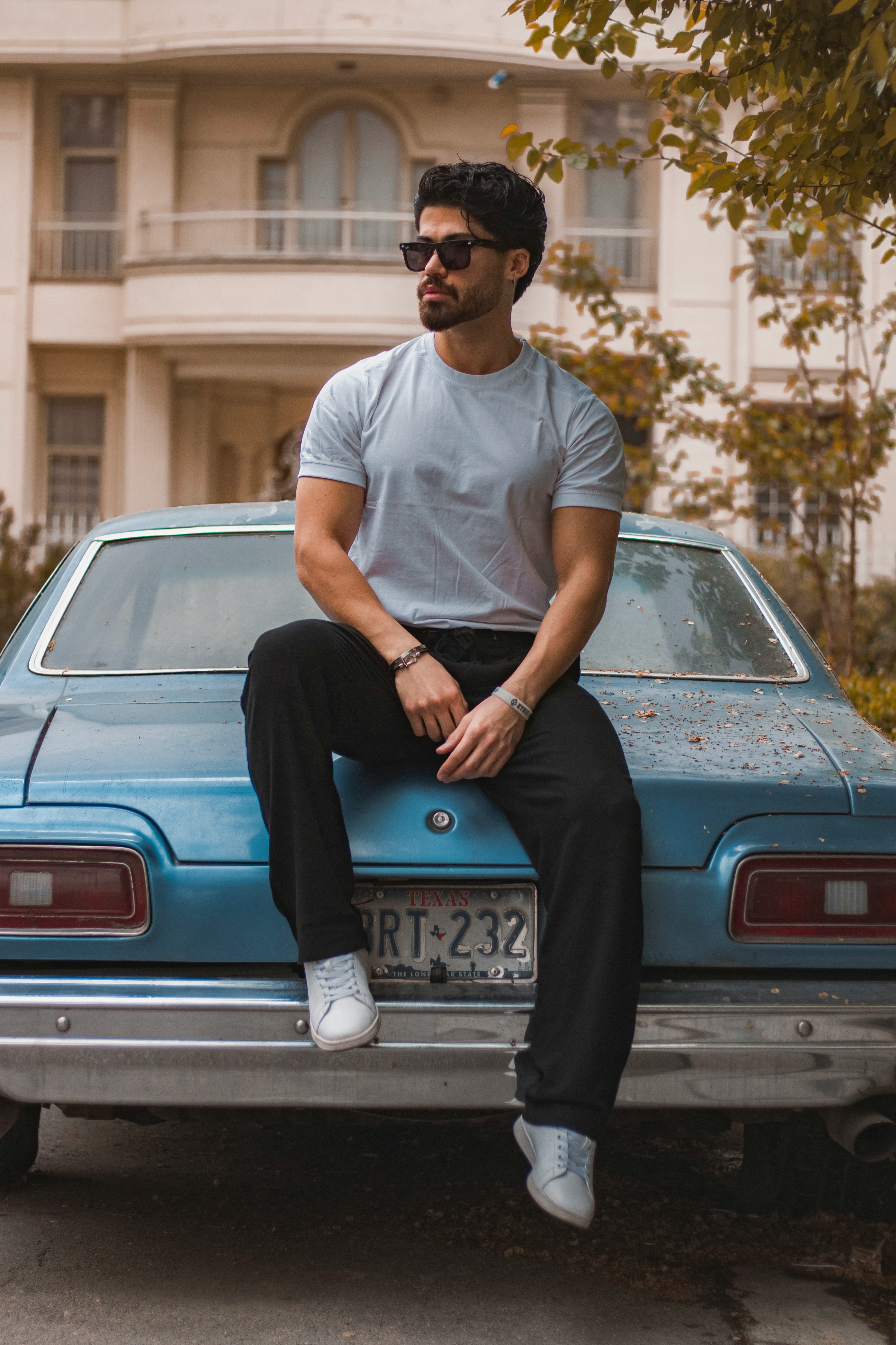 A slightly wider shot of a man in a grey t-shirt and black pants sitting on the back of a weathered blue classic car with a visible license plate.