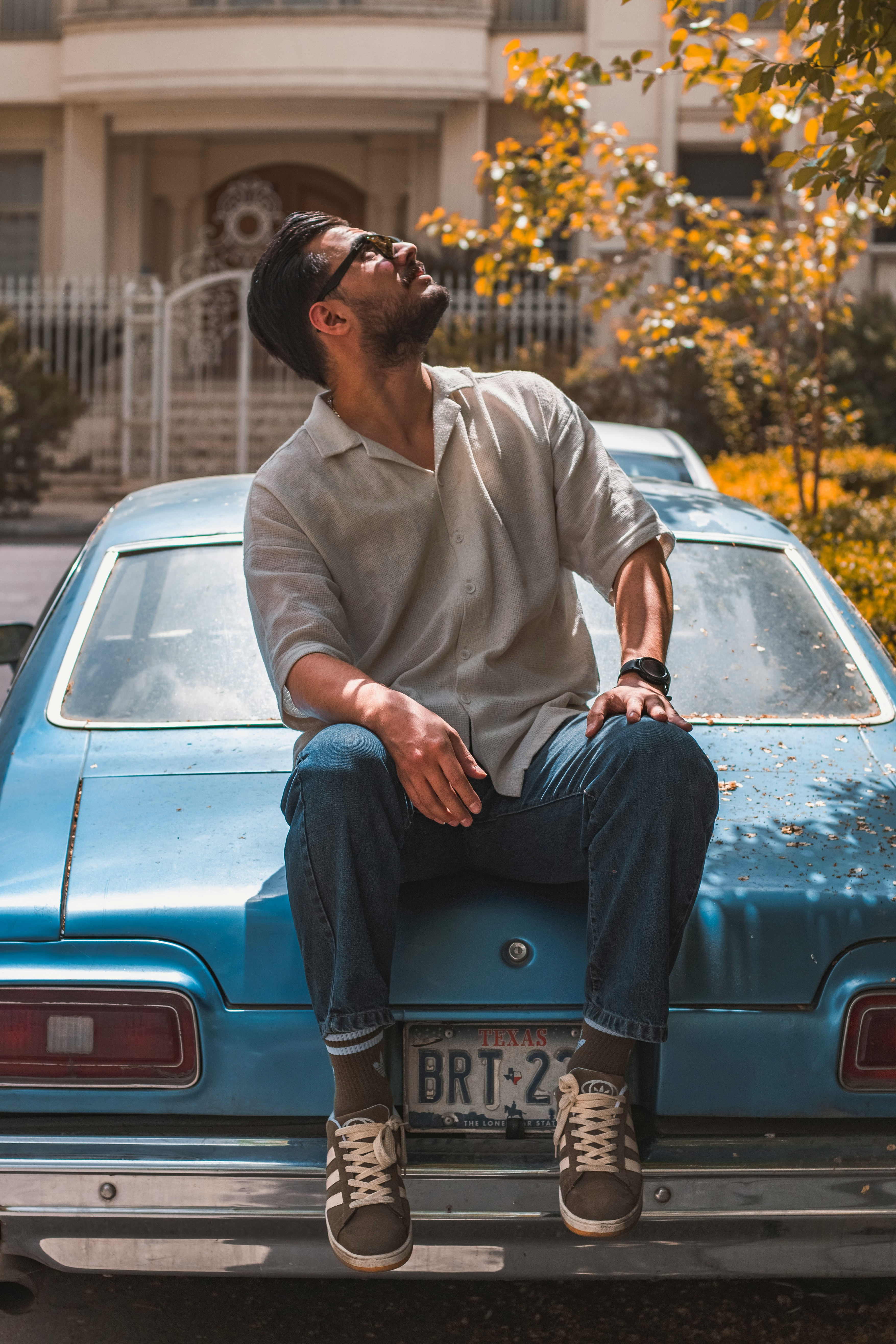 A similar view of a man in a light-colored shirt and jeans leaning against the back of a classic light-blue car with a blurred building in the background.