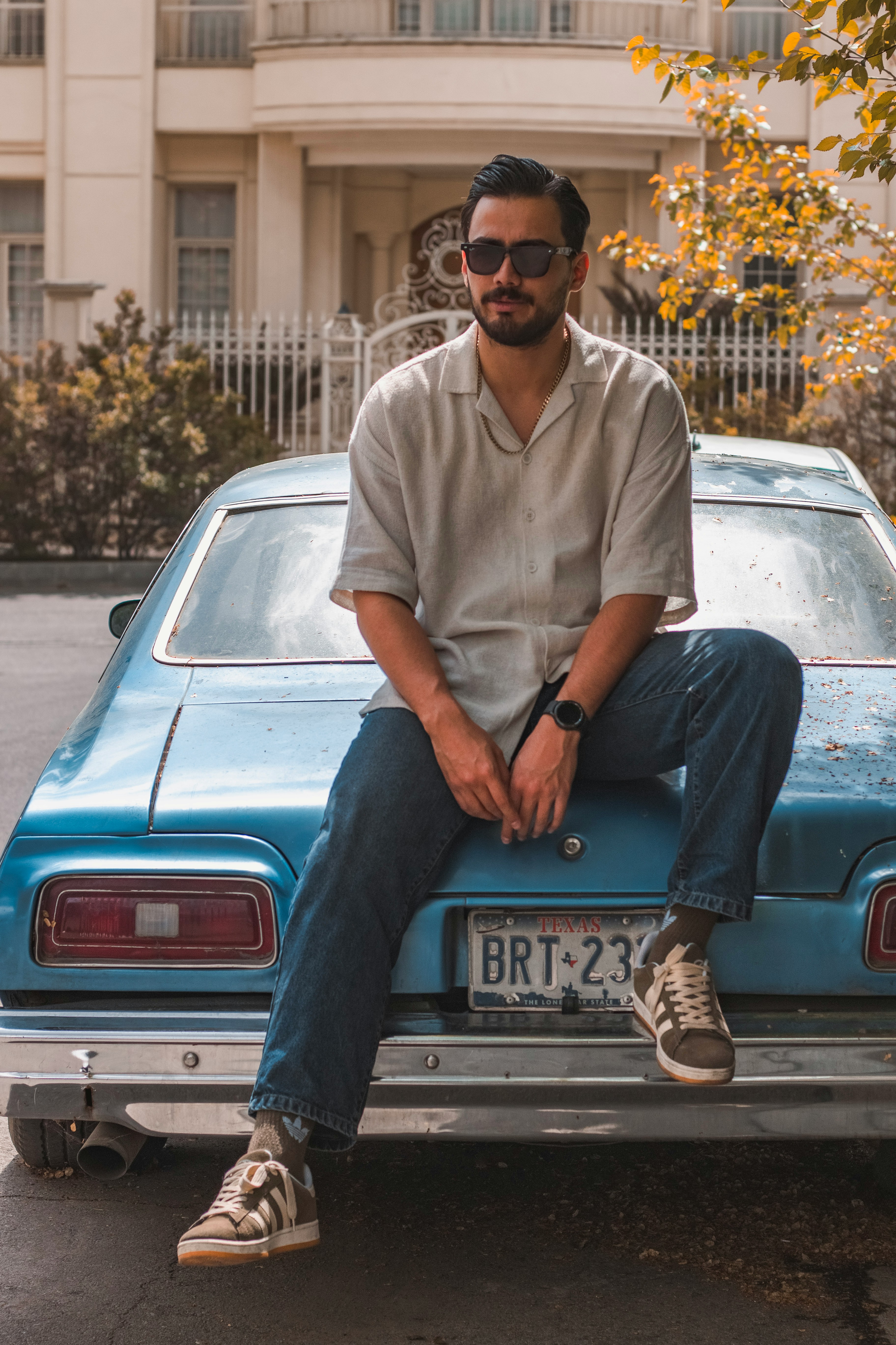 A man in a light grey short-sleeved shirt and jeans sitting on the trunk of a vintage blue car.