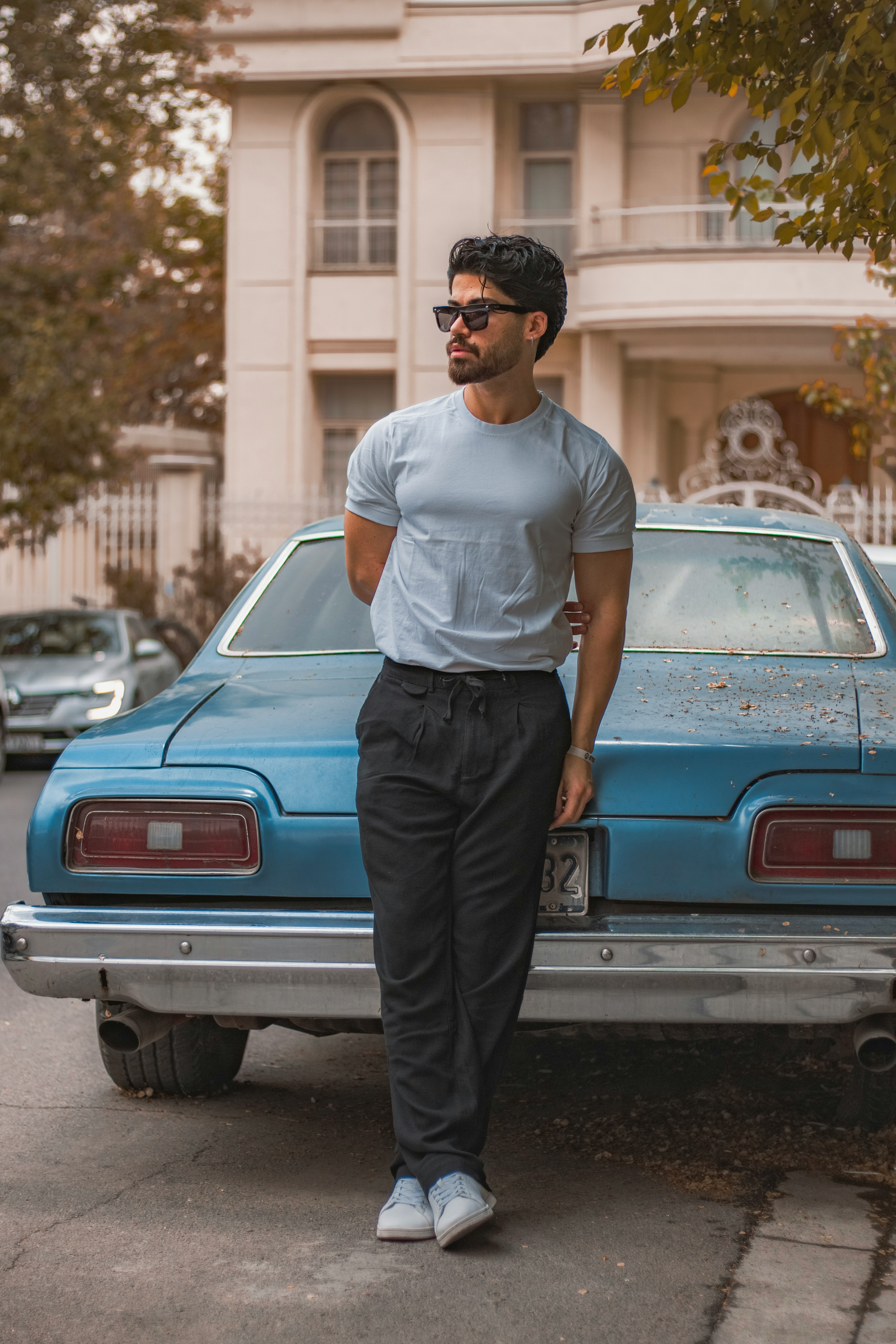 A man in a grey t-shirt and black trousers standing next to the same vintage blue car, leaning against the rear fender.