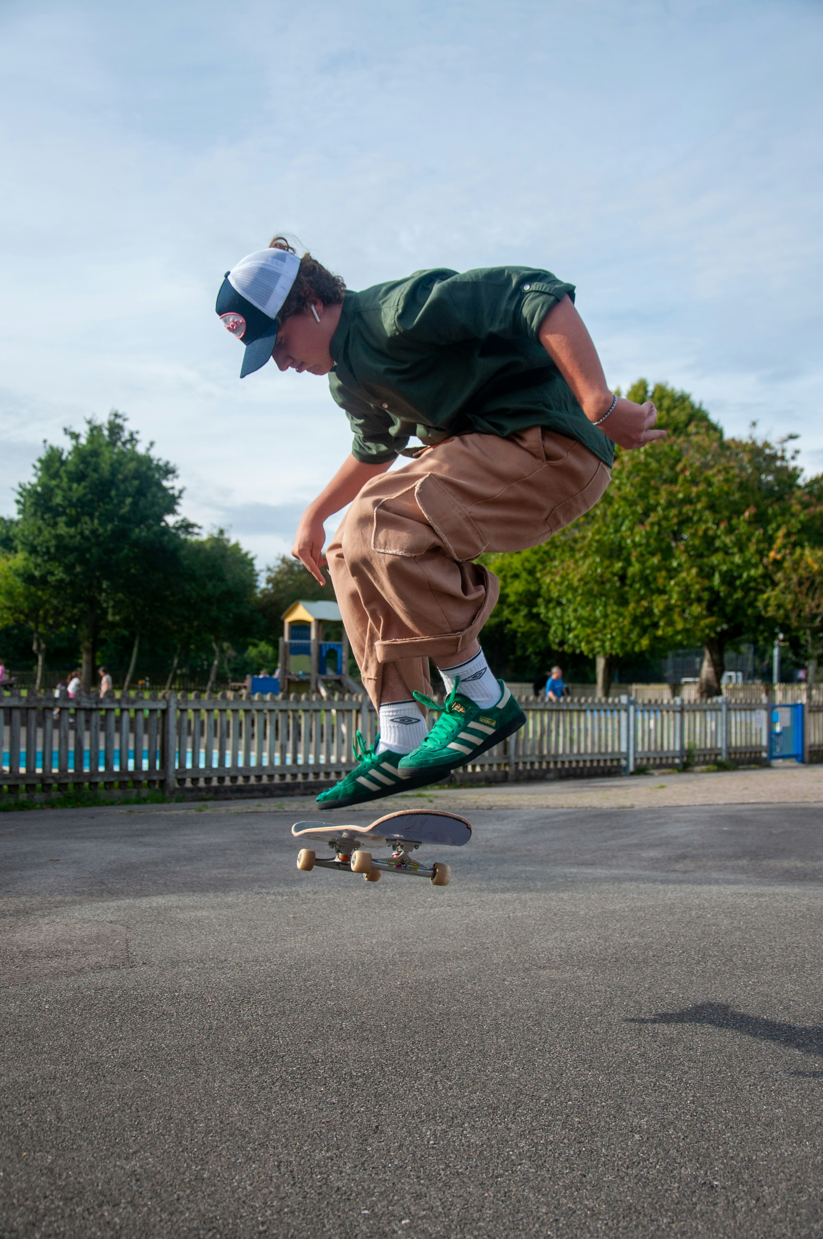 A wide shot of a skateboarder in a green shirt and brown pants captured mid-jump over their board at an outdoor skatepark.