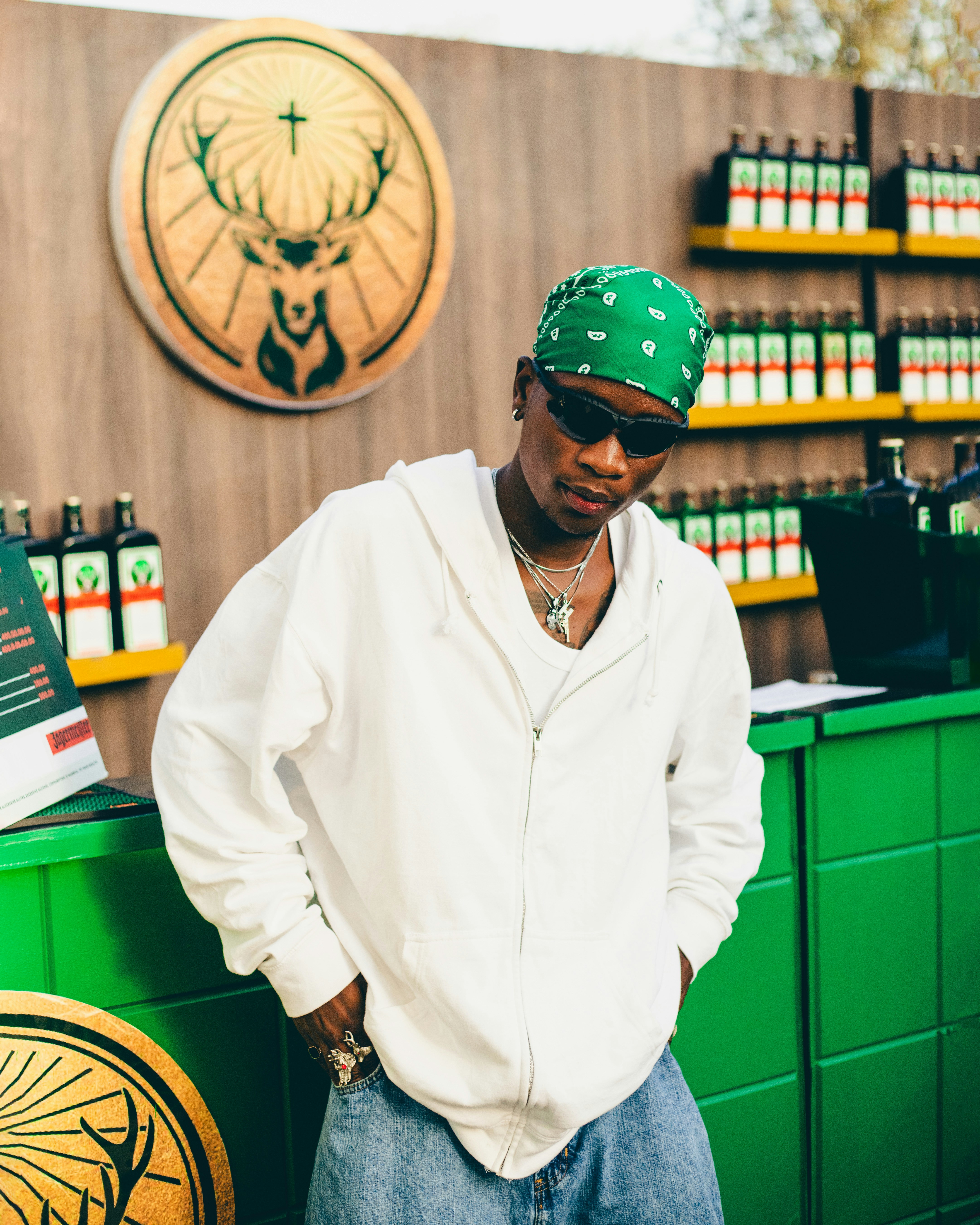 A young man wearing a green patterned bandana, sunglasses, and a white hoodie posing in front of a green bar or stall.