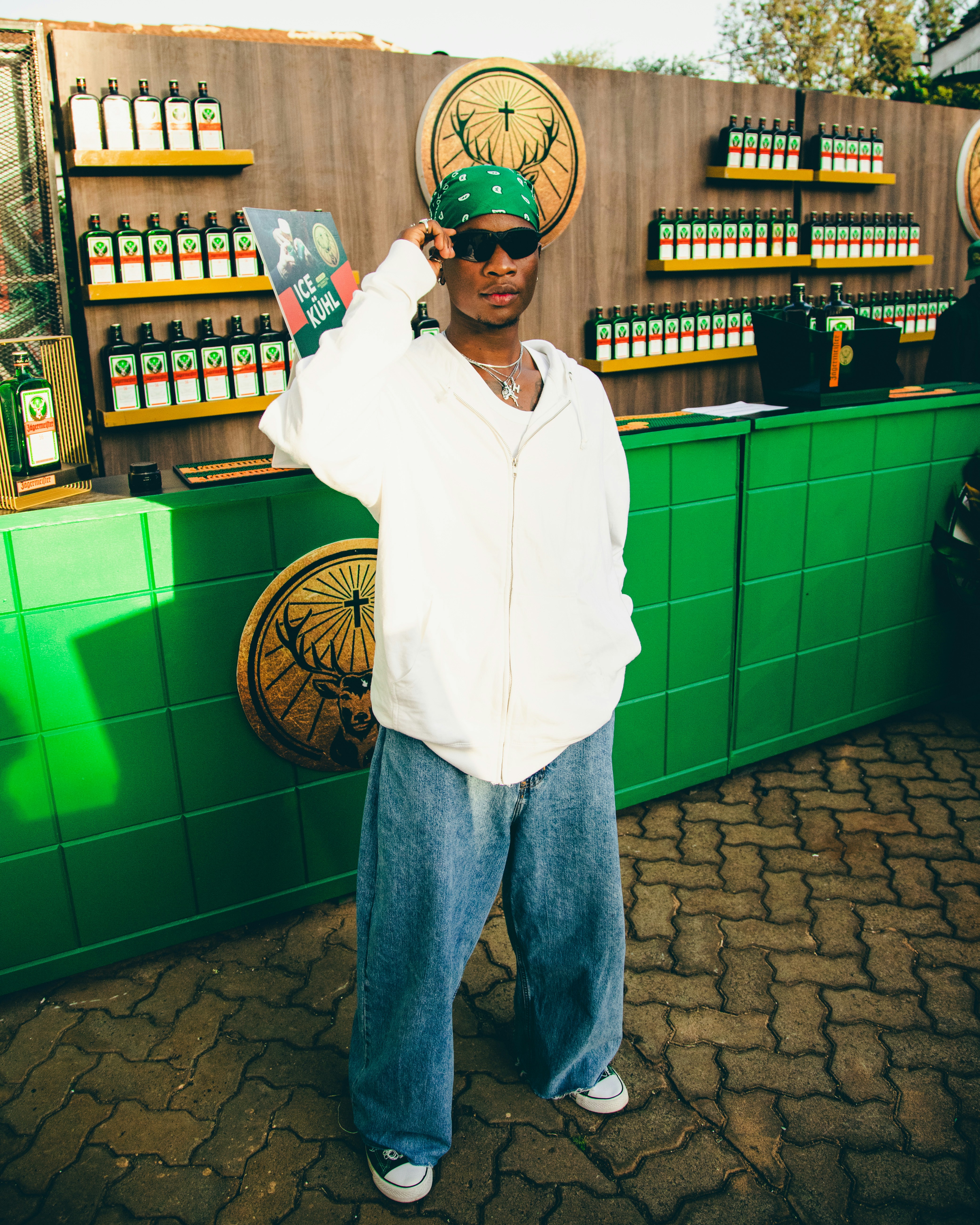 A vertical shot of the young man in the white hoodie and green bandana standing in front of a green counter with bottles of Jägermeister in the background.