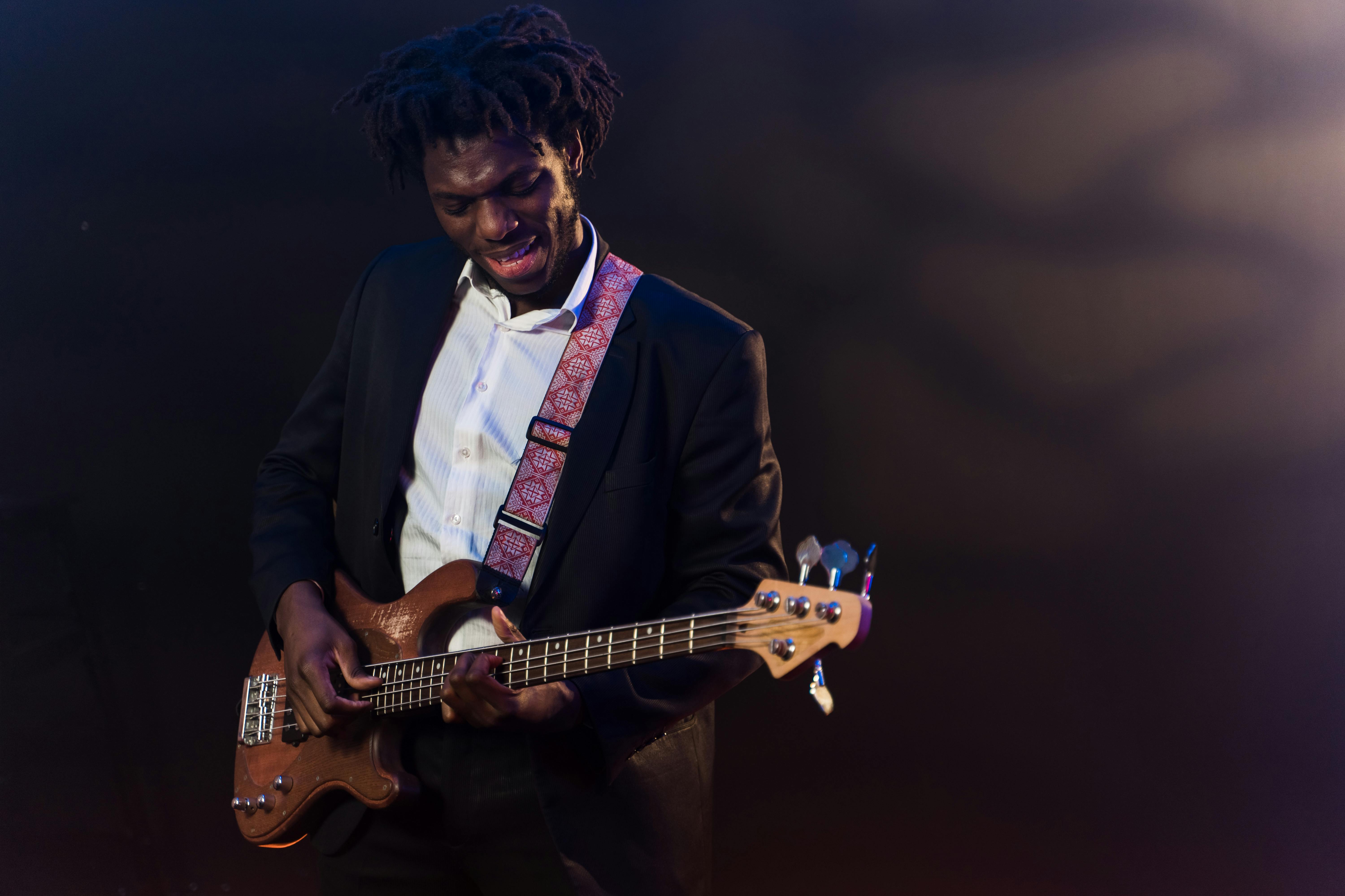 A young Black man in a black suit and white shirt, looking down focused as he plays an electric bass guitar with a patterned strap.