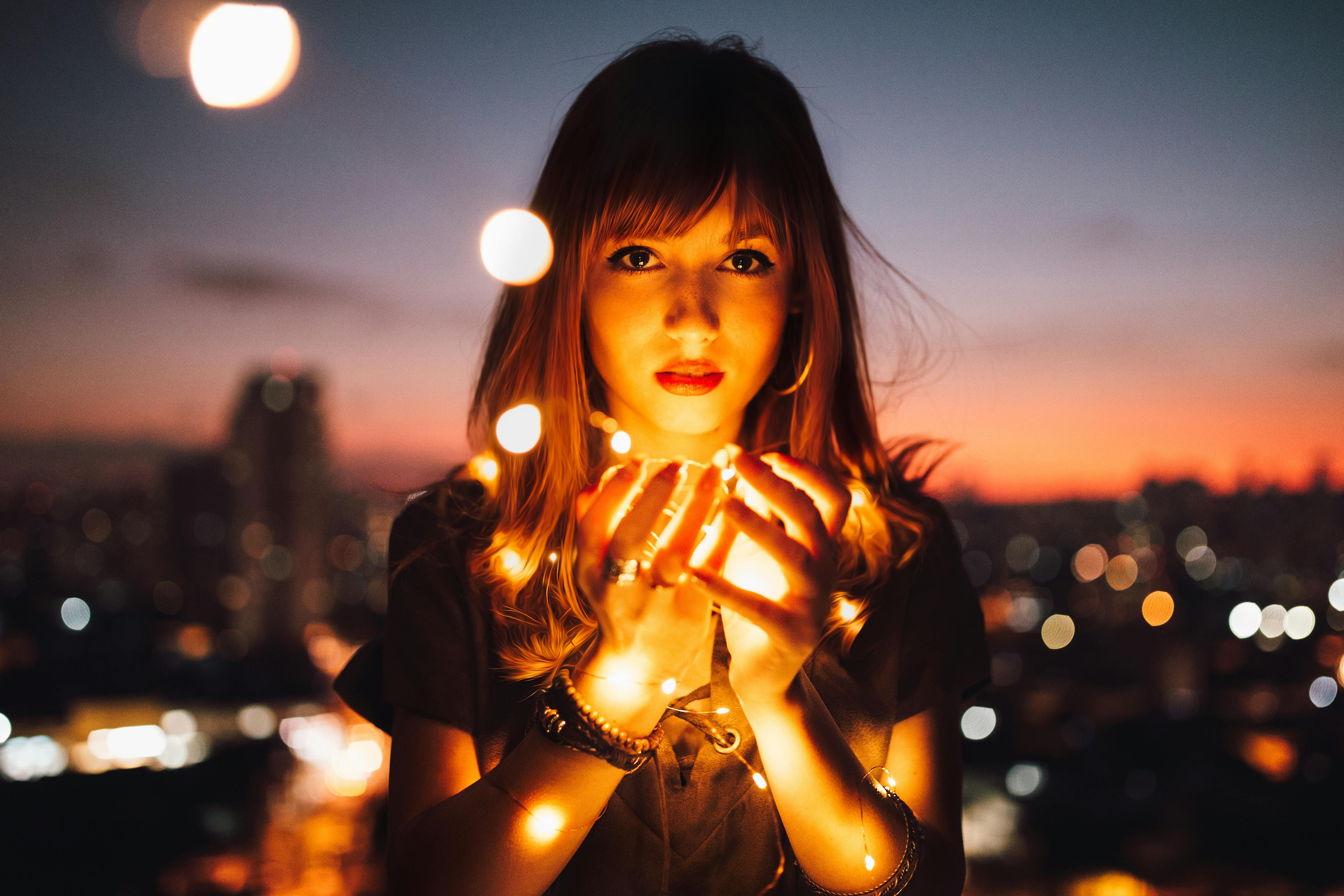 A portrait of a young woman with bangs holding glowing fairy lights close to her face, with a blurred city skyline at dusk in the background.