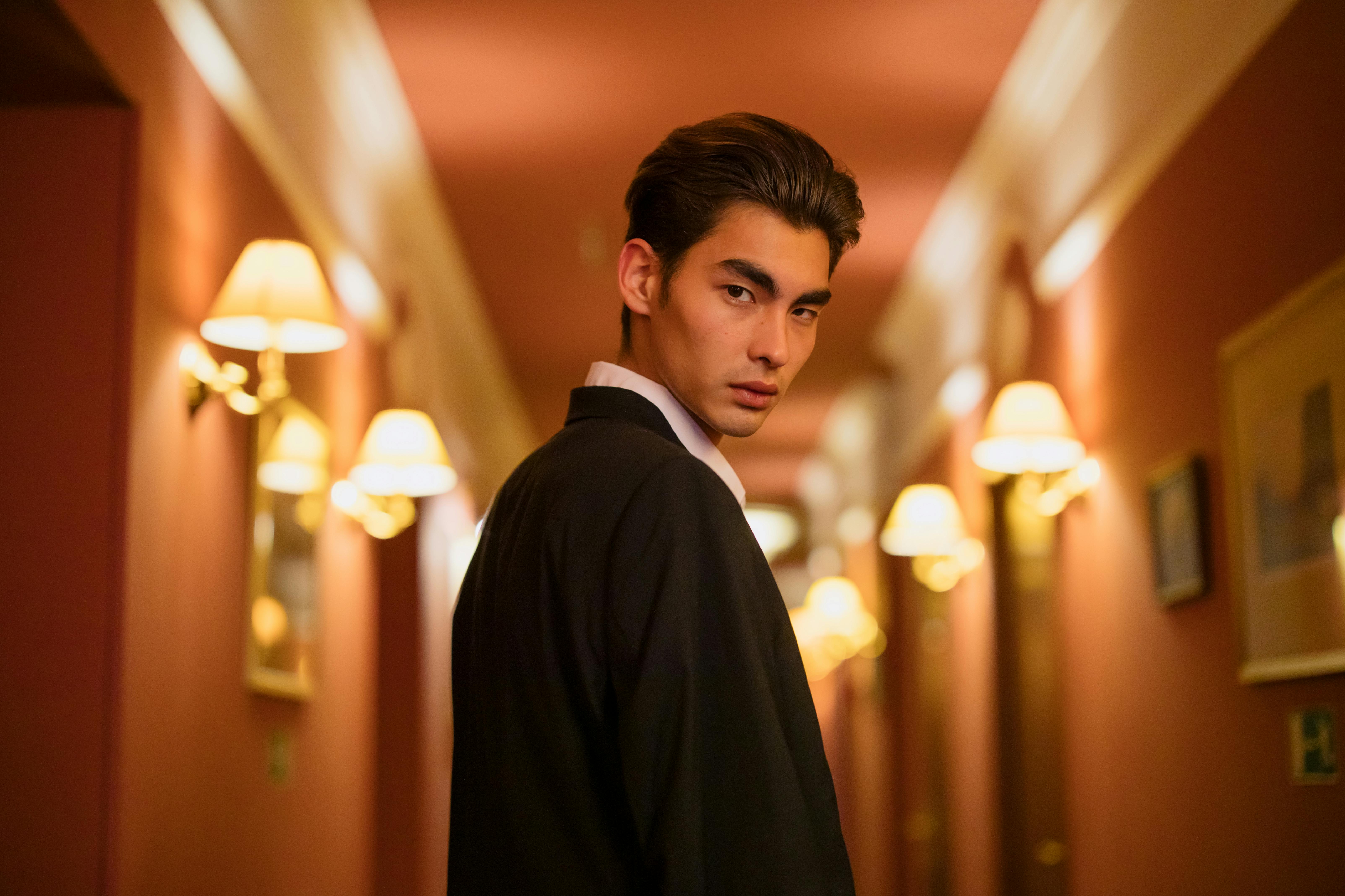 A young man in a black suit looking over his shoulder in a warm, elegantly lit hallway with classical wall lamps.