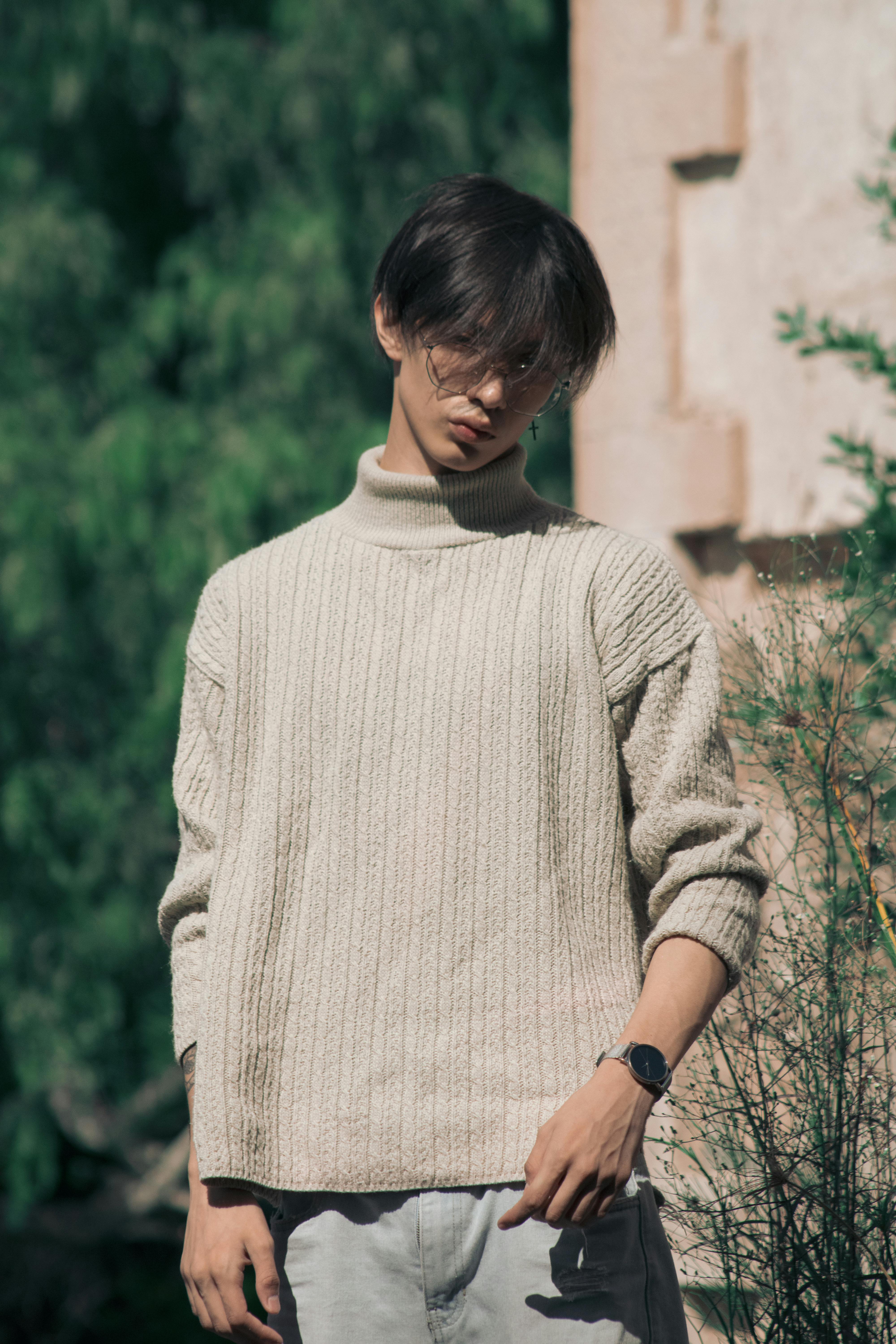 A young man wearing a chunky, cream-colored knitted turtleneck sweater, posing outdoors against a soft green background.