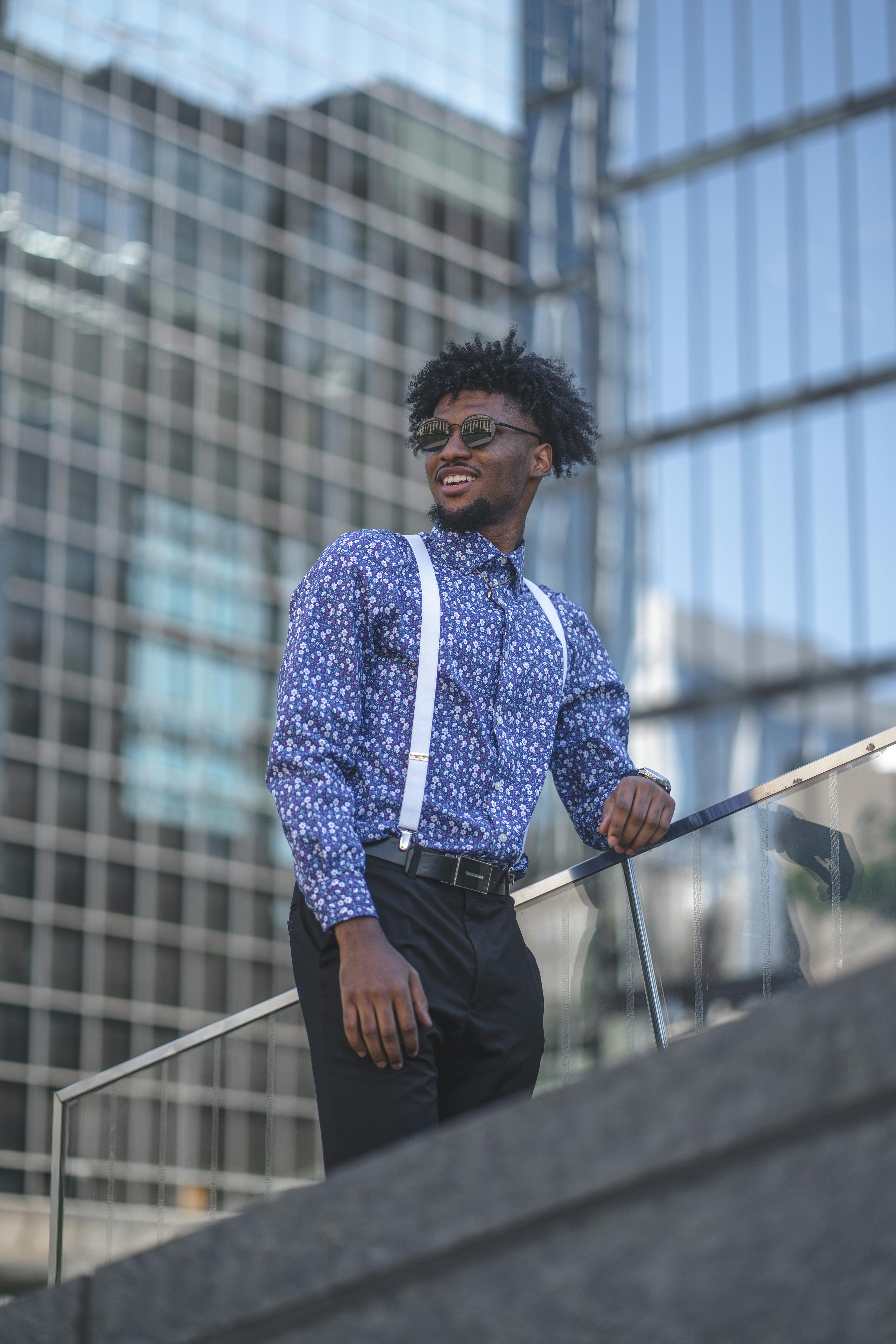 A smiling Black man in a blue floral patterned shirt and white suspenders, leaning against a railing in a bright urban setting.