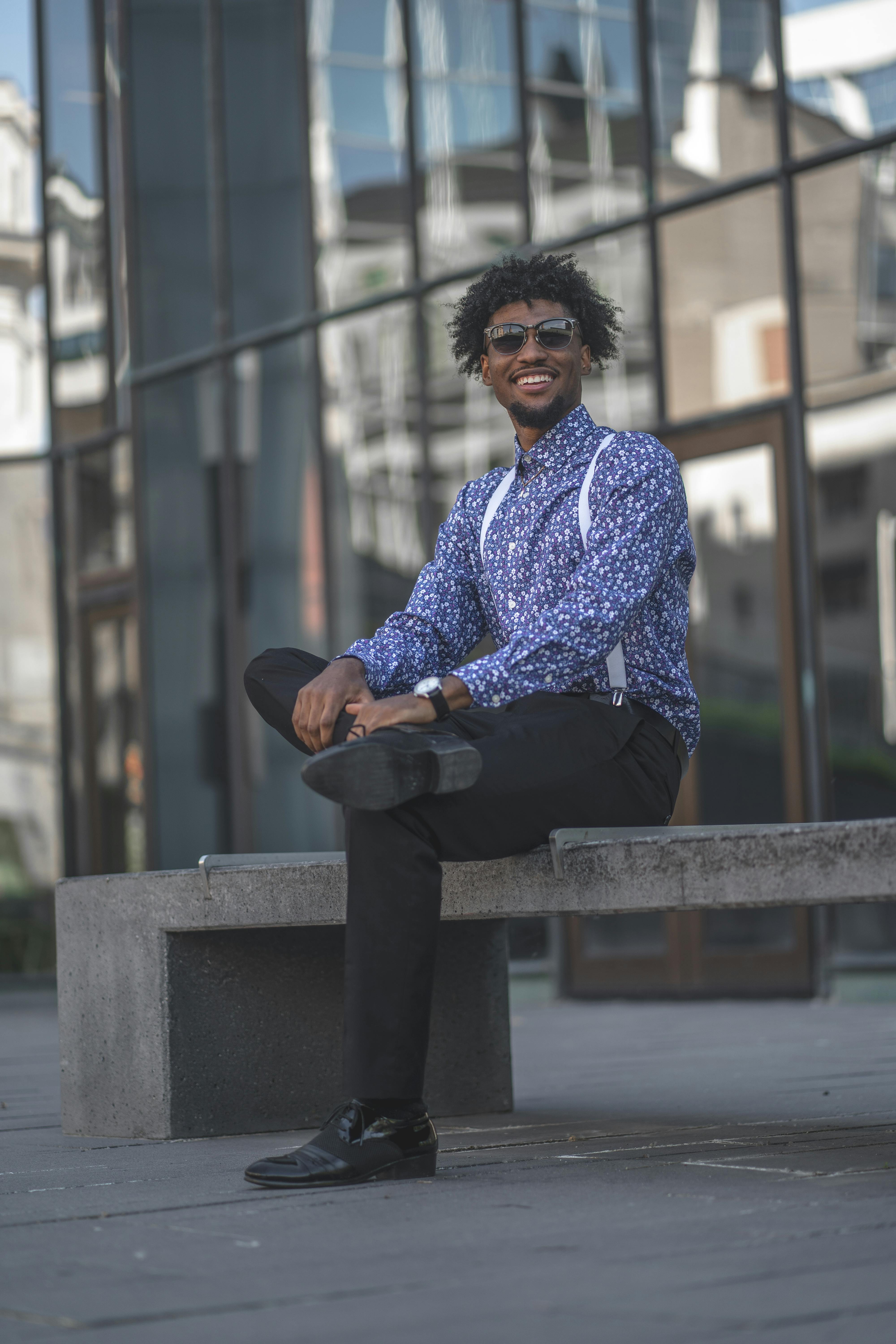 A candid, full-body shot of the same man in the floral shirt and suspenders, laughing while sitting on a stone ledge in the city.