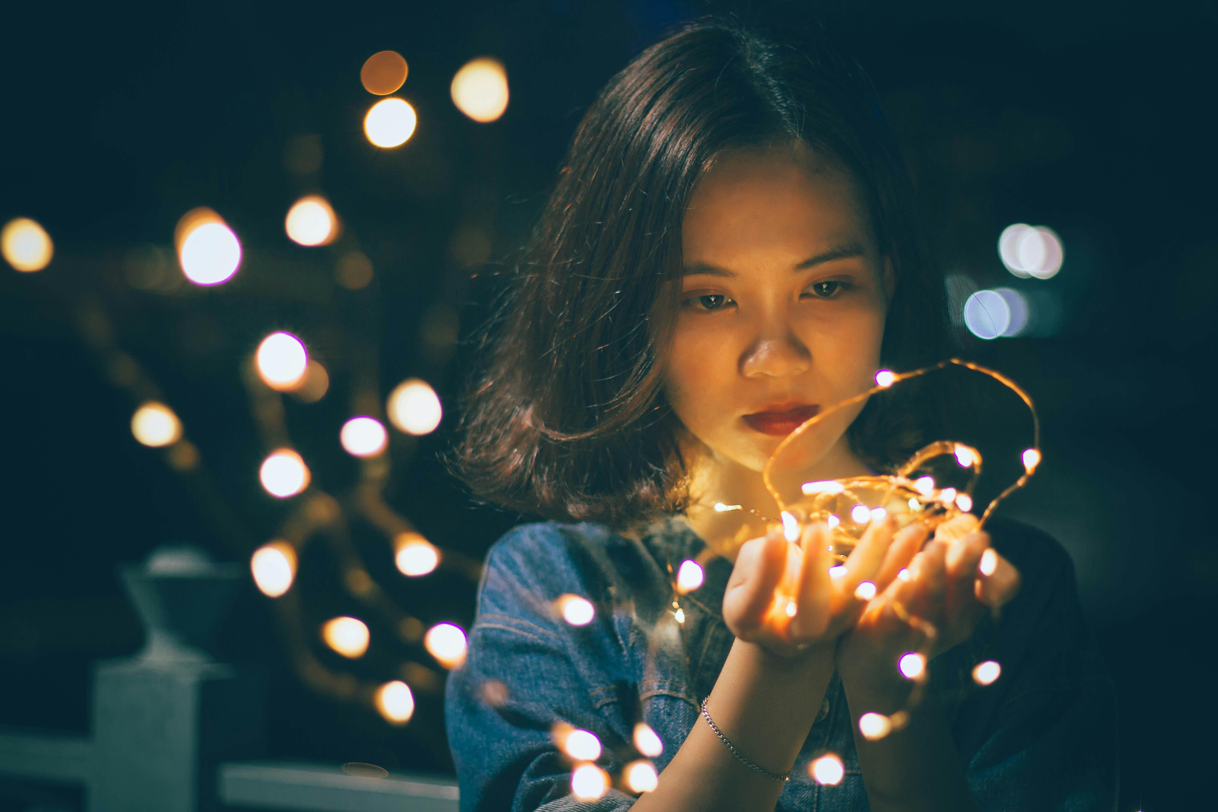 A young woman with shoulder-length dark hair holding a bundle of glowing copper fairy lights in her hands, looking at them with a soft, focused expression.