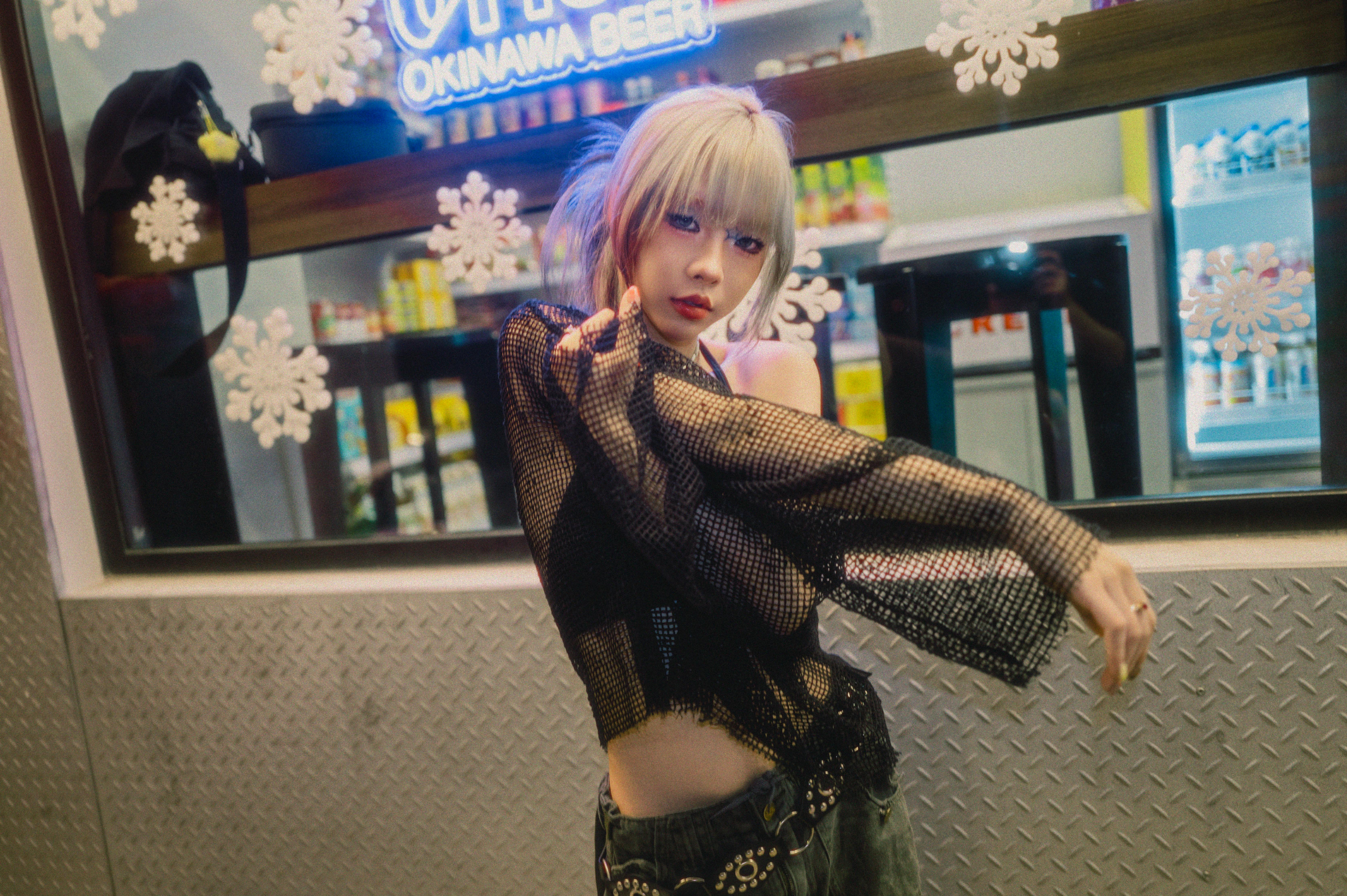 A young woman with short blonde hair and a dark mesh top posing in front of an "Okinawa Beer" sign with snowflakes, creating a cool, urban vibe.