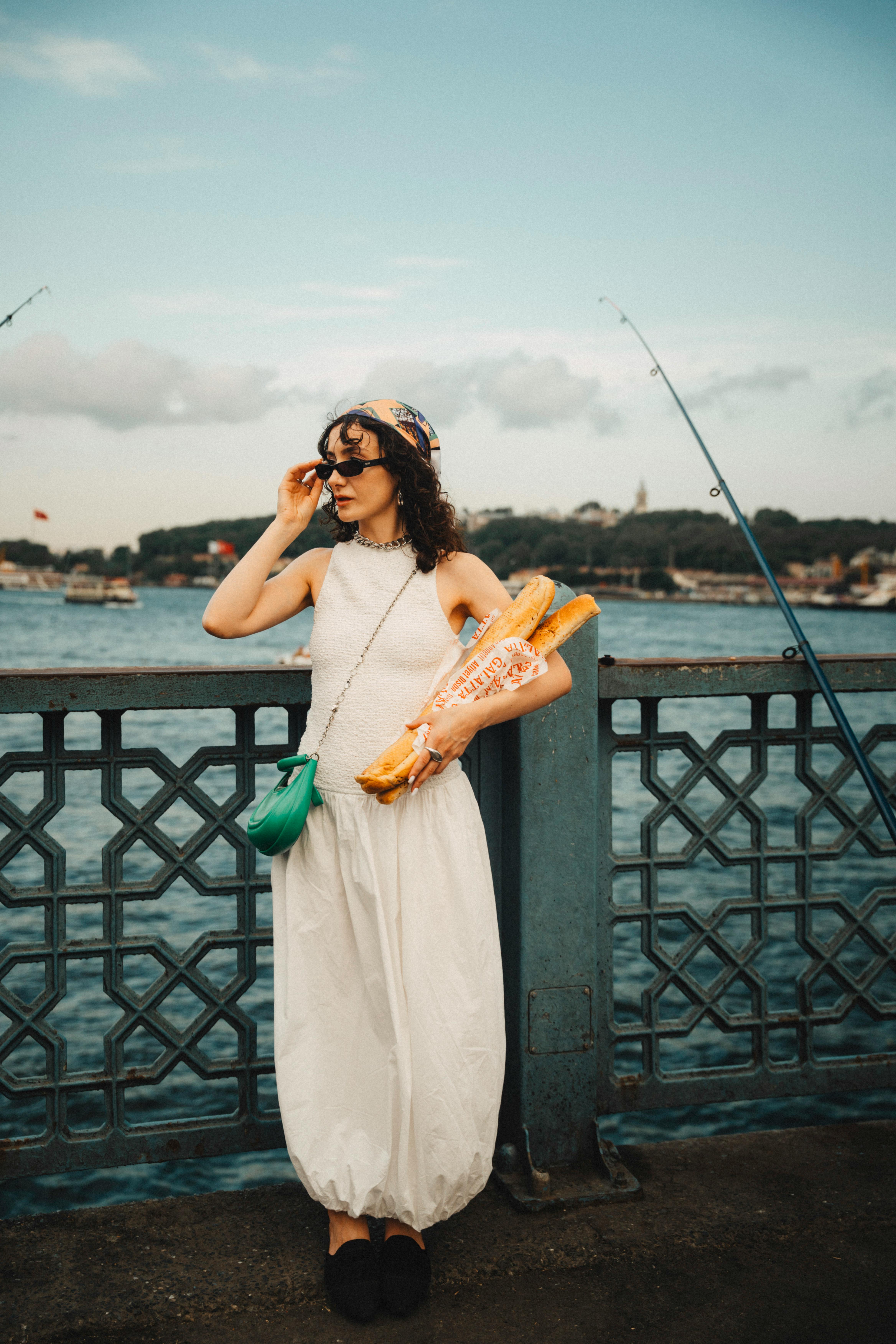 A woman in a white dress, sunglasses, and a colorful headscarf leaning against a railing by the water, holding two long baguettes.