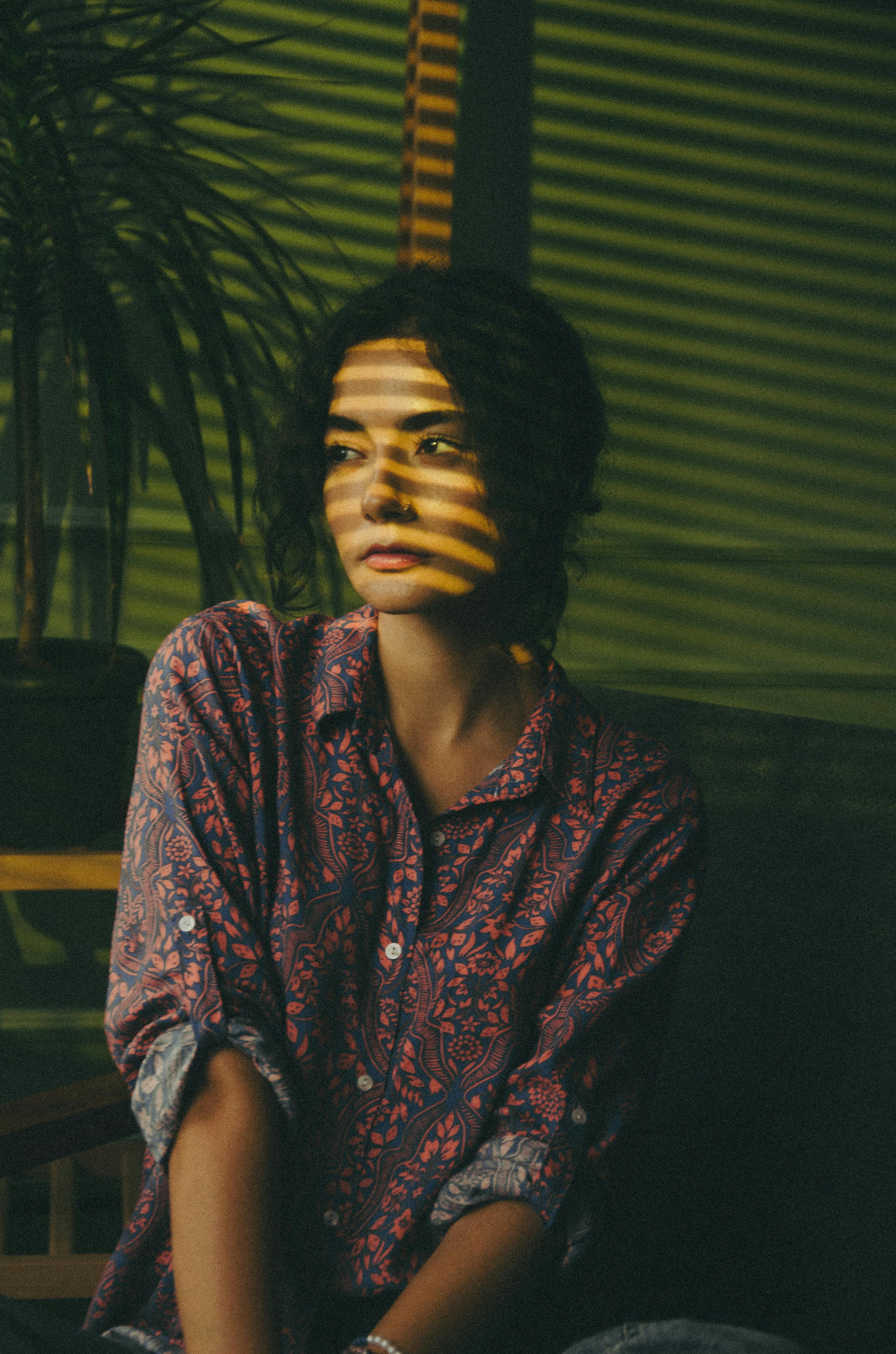 A moody portrait of a woman in a patterned red shirt, with sunlight filtering through window blinds to cast horizontal shadow stripes across her face.