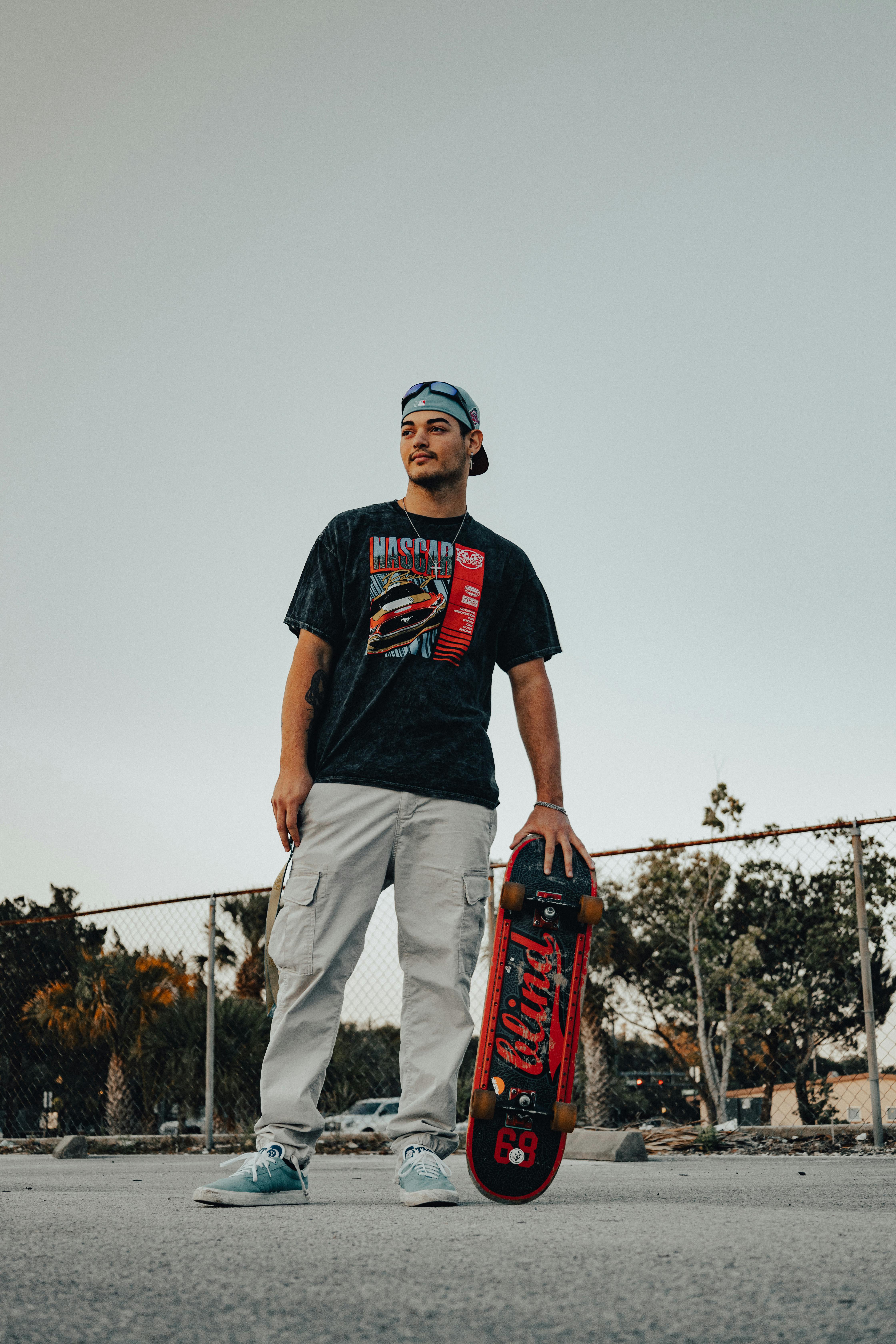 A low-angle shot of a young man in a graphic t-shirt and grey cargo pants standing outdoors, holding a skateboard by his side.
