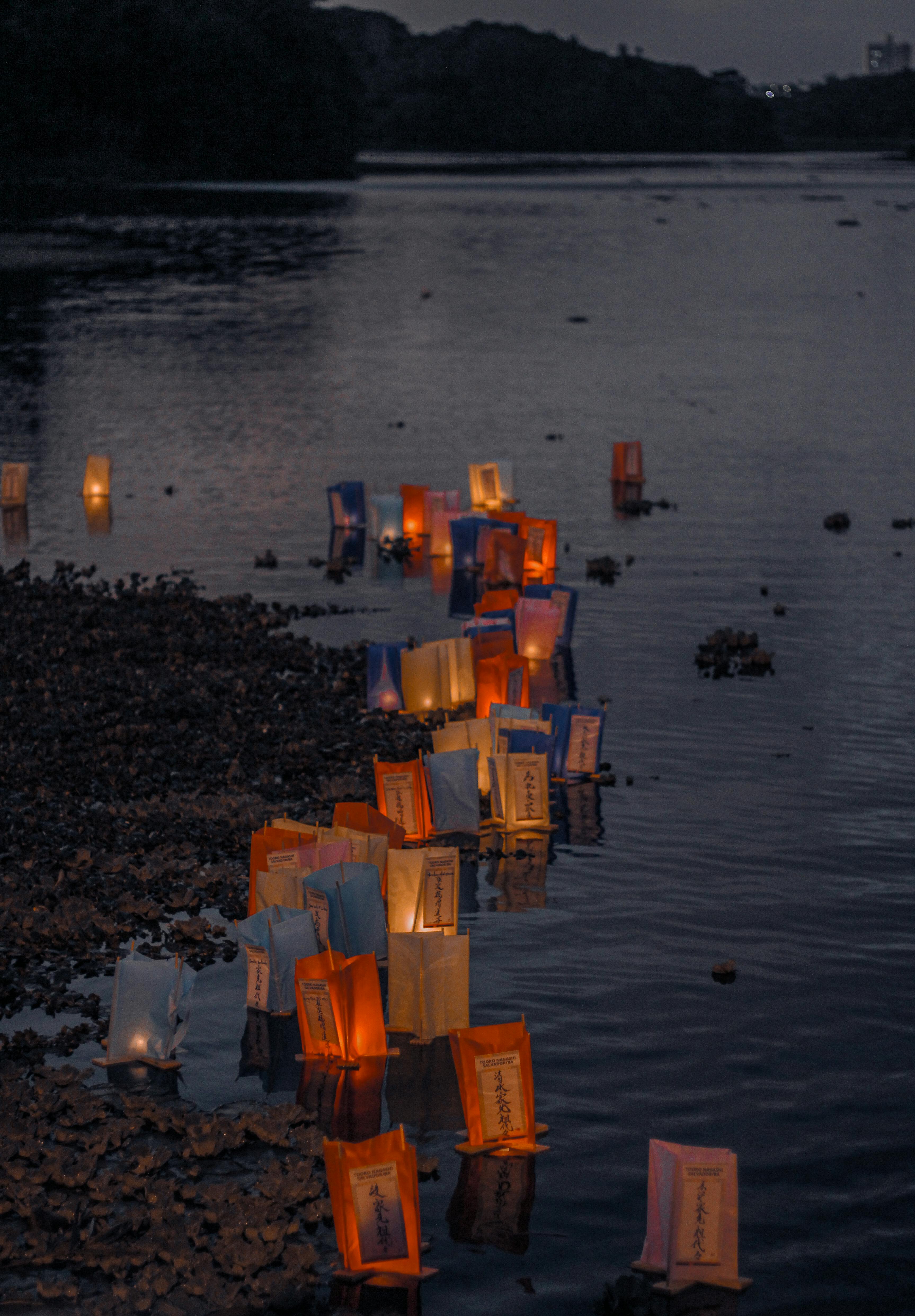 Dozens of glowing orange and blue paper lanterns floating on a dark body of water at dusk