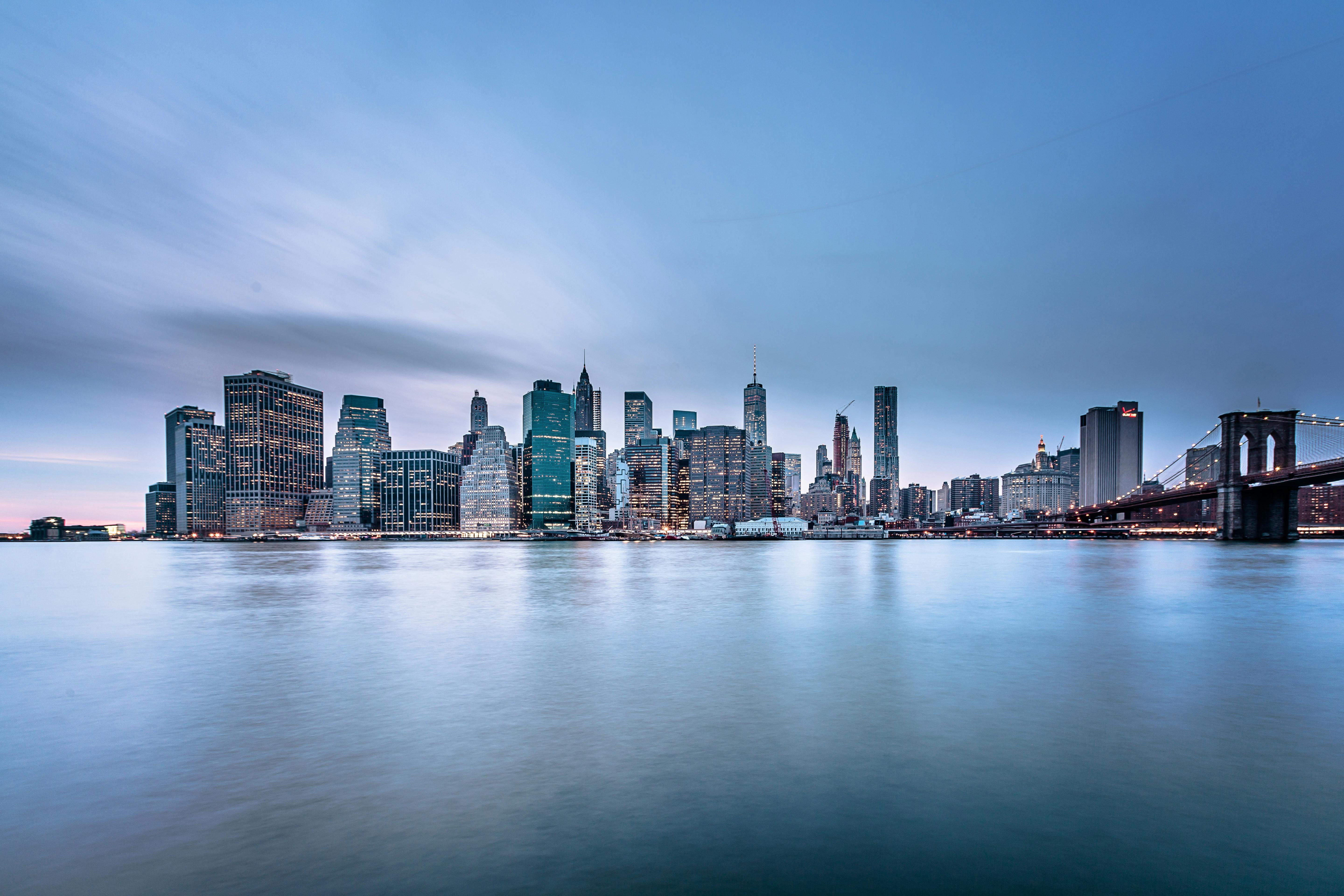 Lower Manhattan skyline, including One World Trade Center, seen across the water under a dramatic, cloudy sky.