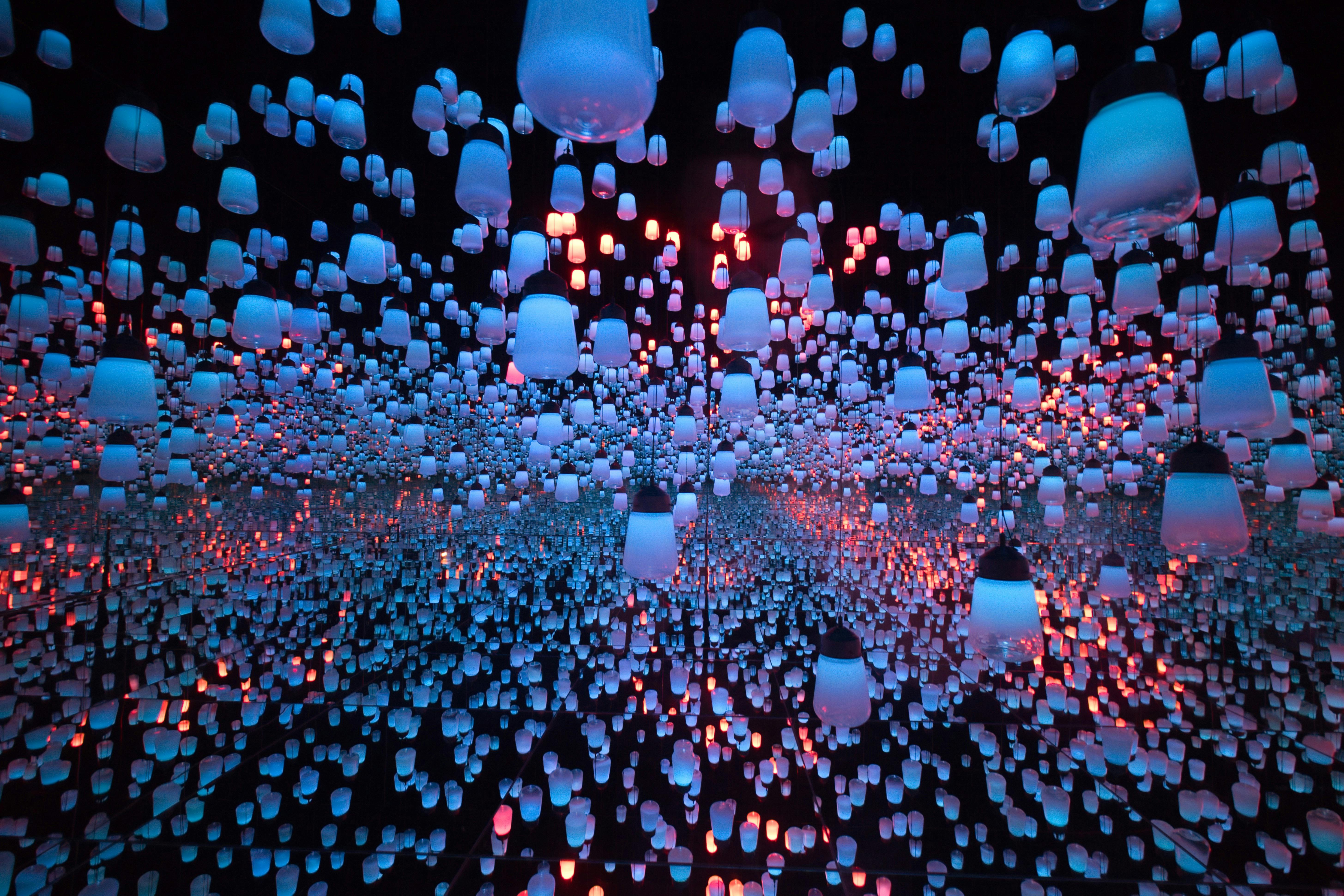 An immersive art installation featuring hundreds of glowing blue and purple lanterns suspended in a dark, mirrored room, creating an infinite field of light.