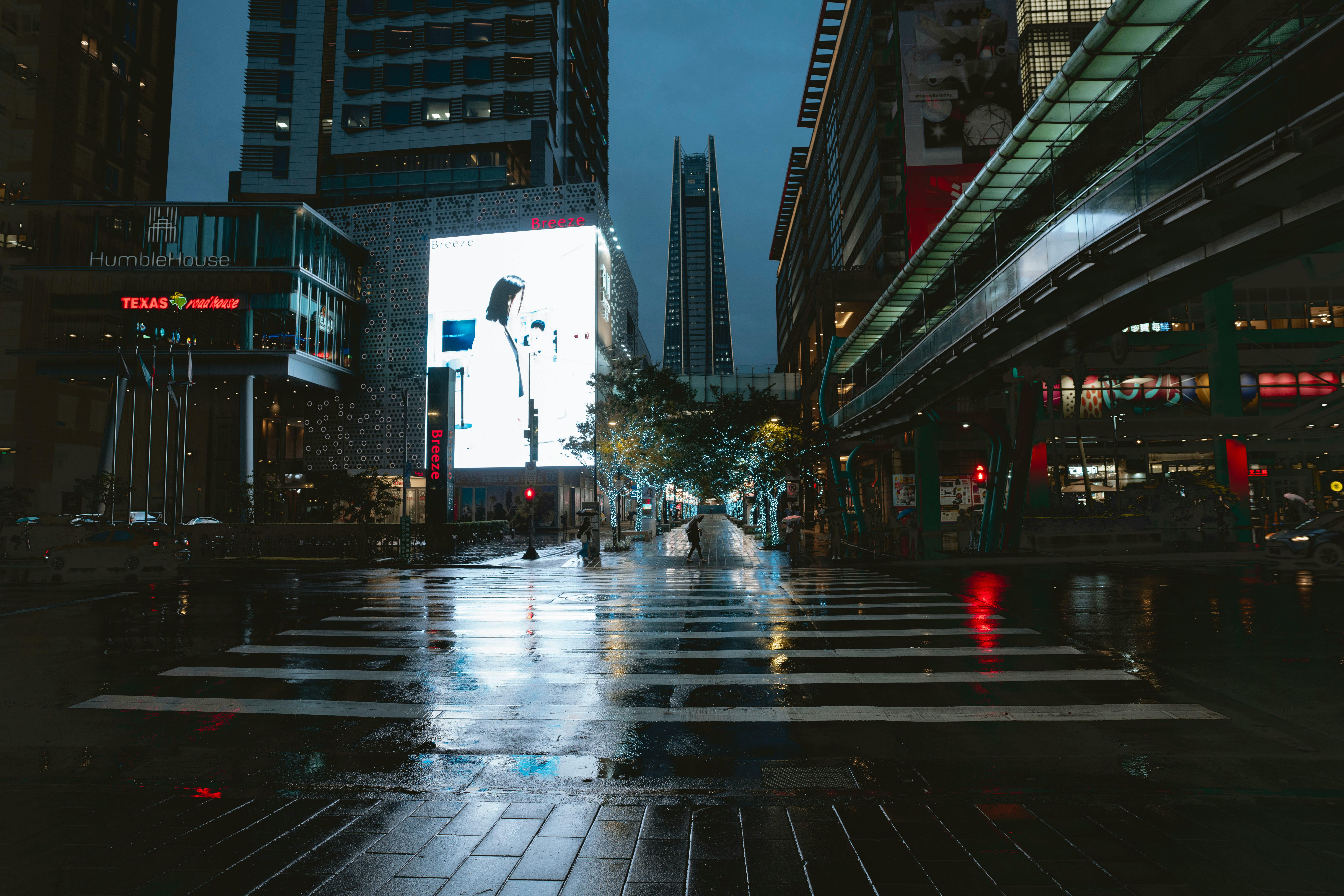 A wide, wet city street at night featuring a bright, glowing billboard and a pedestrian crosswalk reflecting city lights.