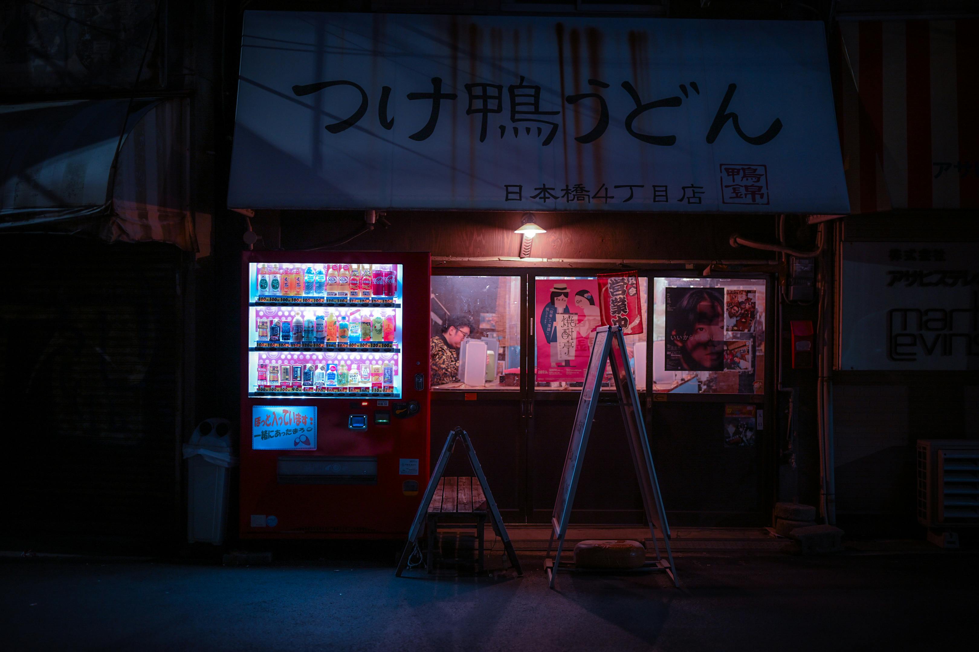 Traditional Japanese storefront at night with glowing vending machines and posters, under a sign for "Tsukamo Udon."