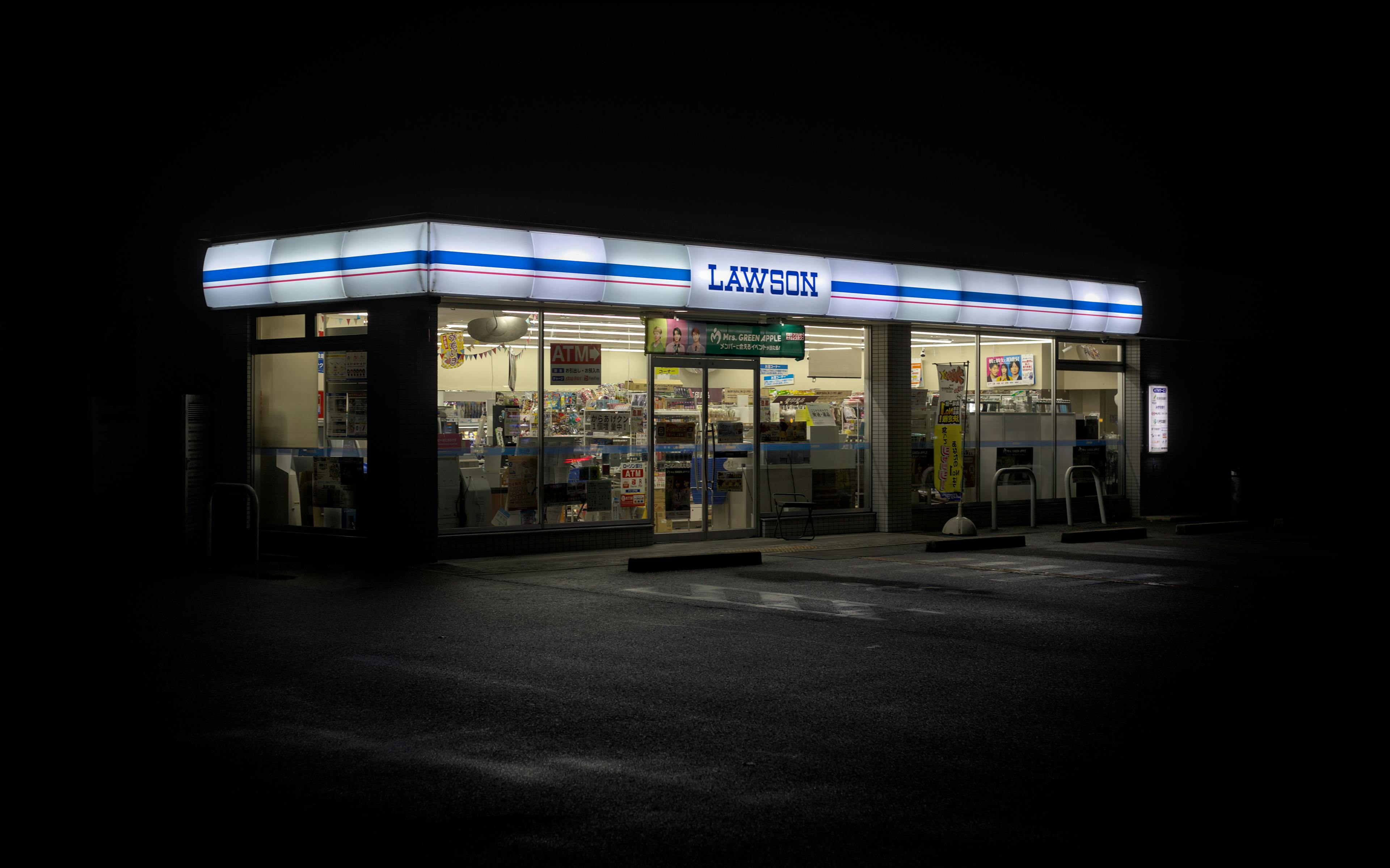 Lawson convenience store standing brightly lit in the middle of a dark, empty parking lot at night.