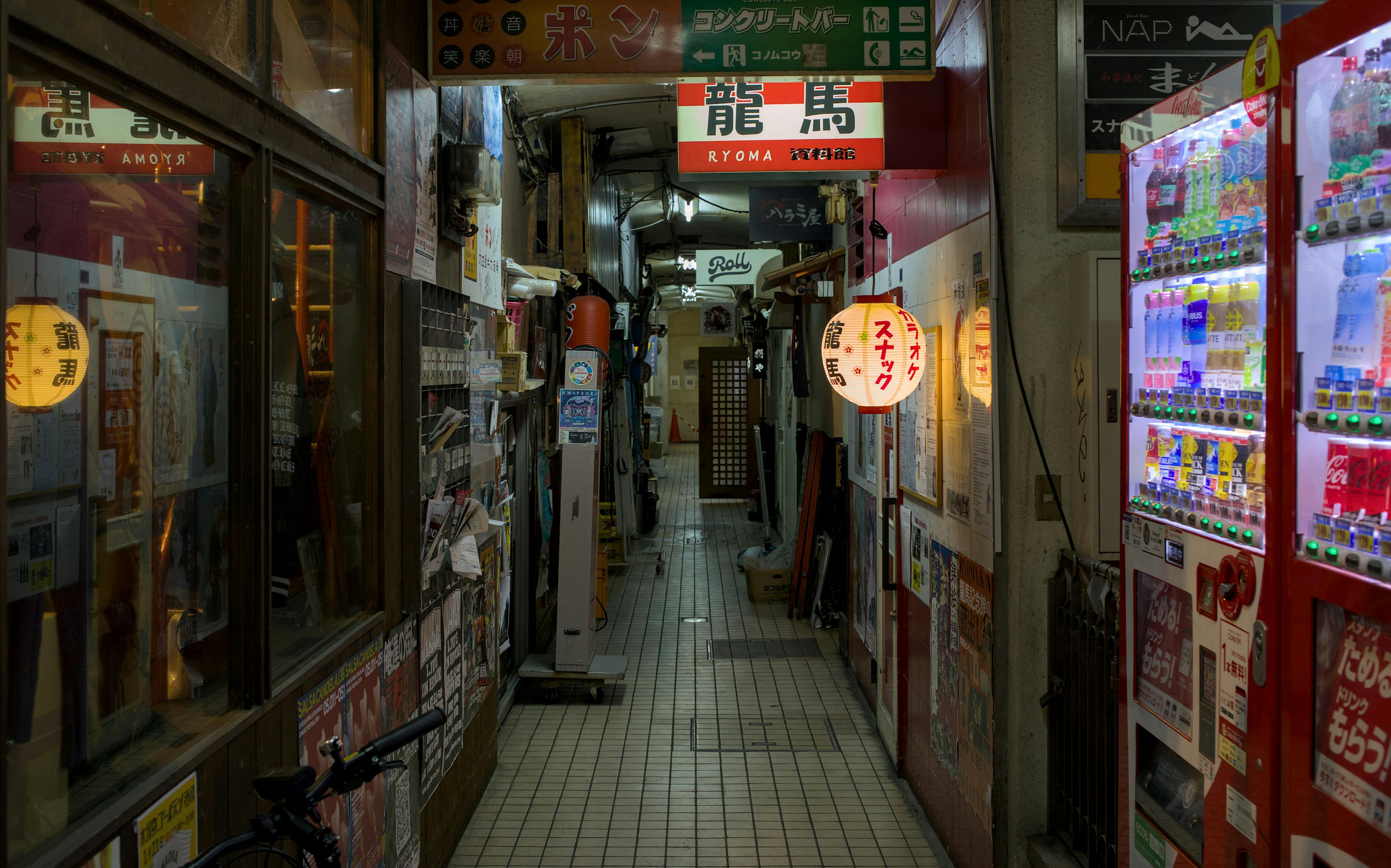 A narrow, cluttered Japanese alleyway (yokocho) at night, filled with small shop entrances, glowing signs, and a vending machine.