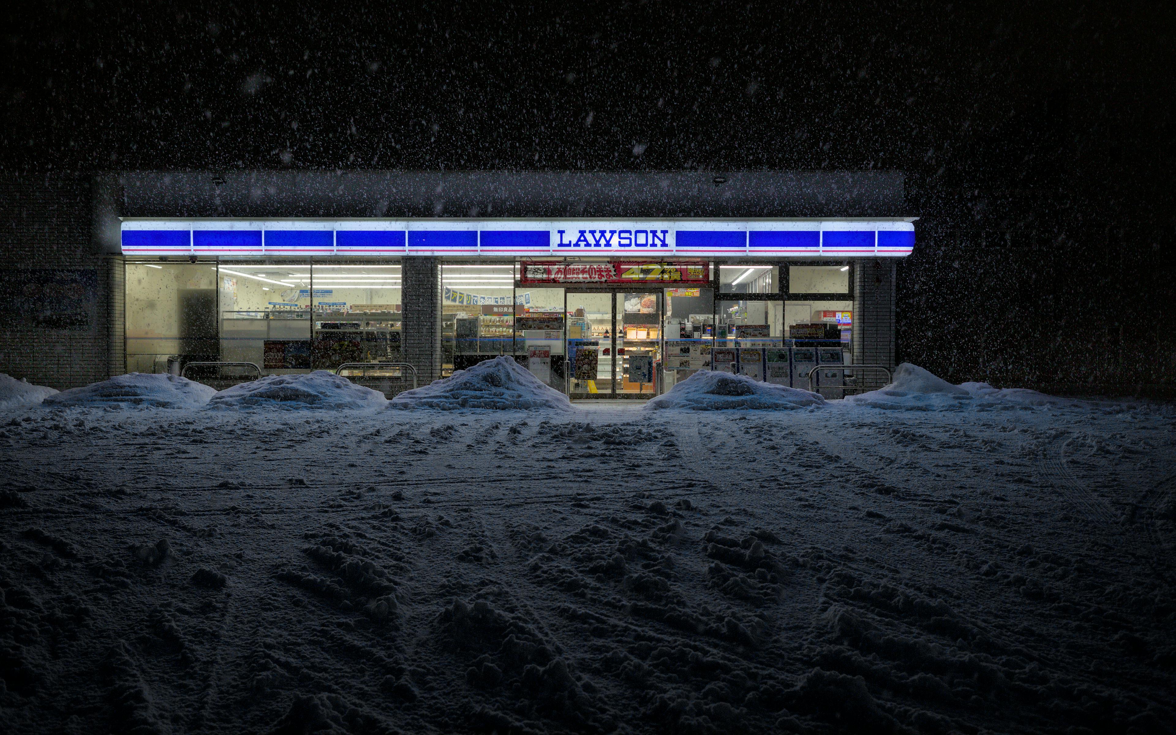 A Lawson convenience store glowing at night, surrounded by deep, heavy snow during a winter storm.