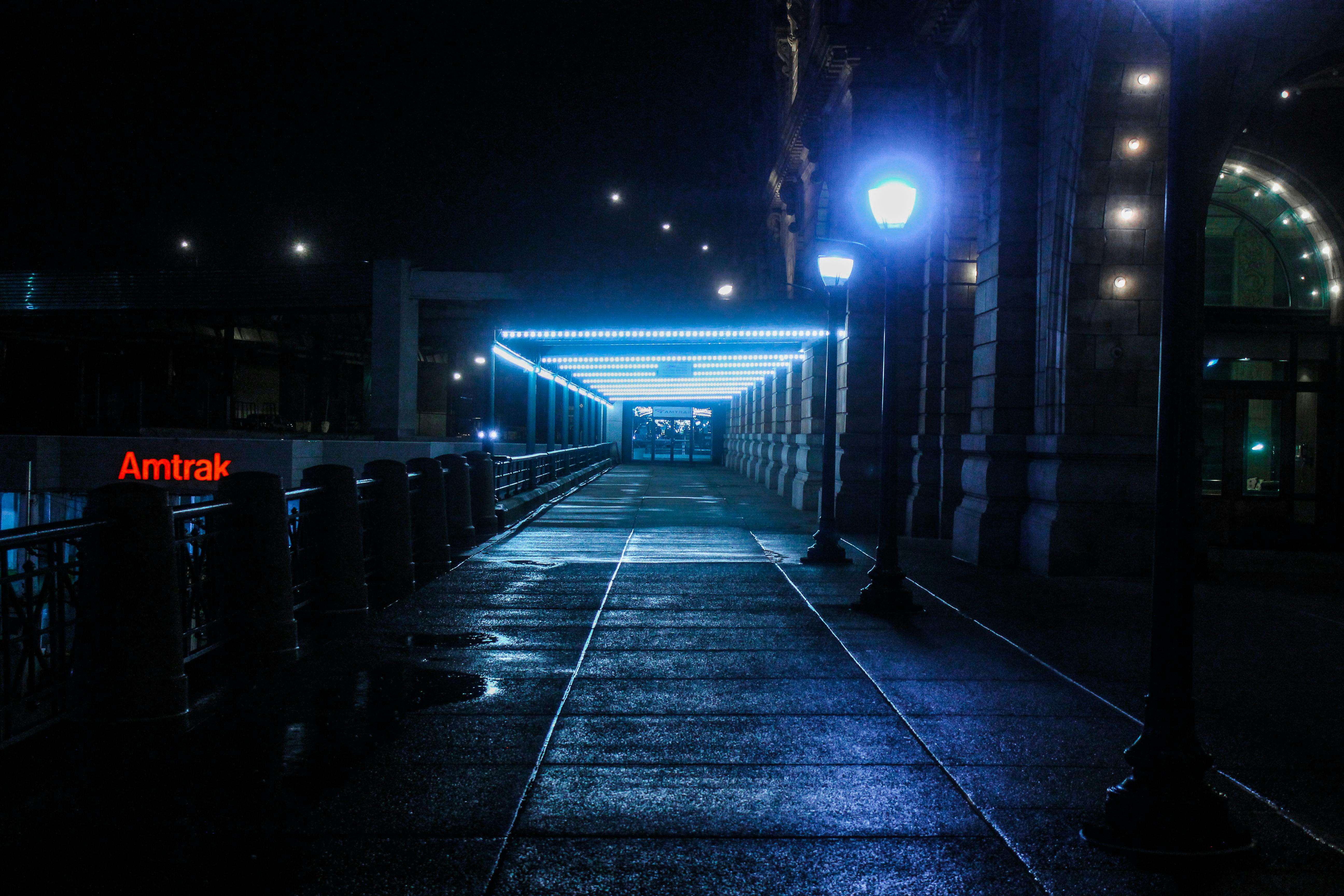 A dark, wet Amtrak station walkway at night, illuminated by blue neon tunnel lights and distant street lamps.