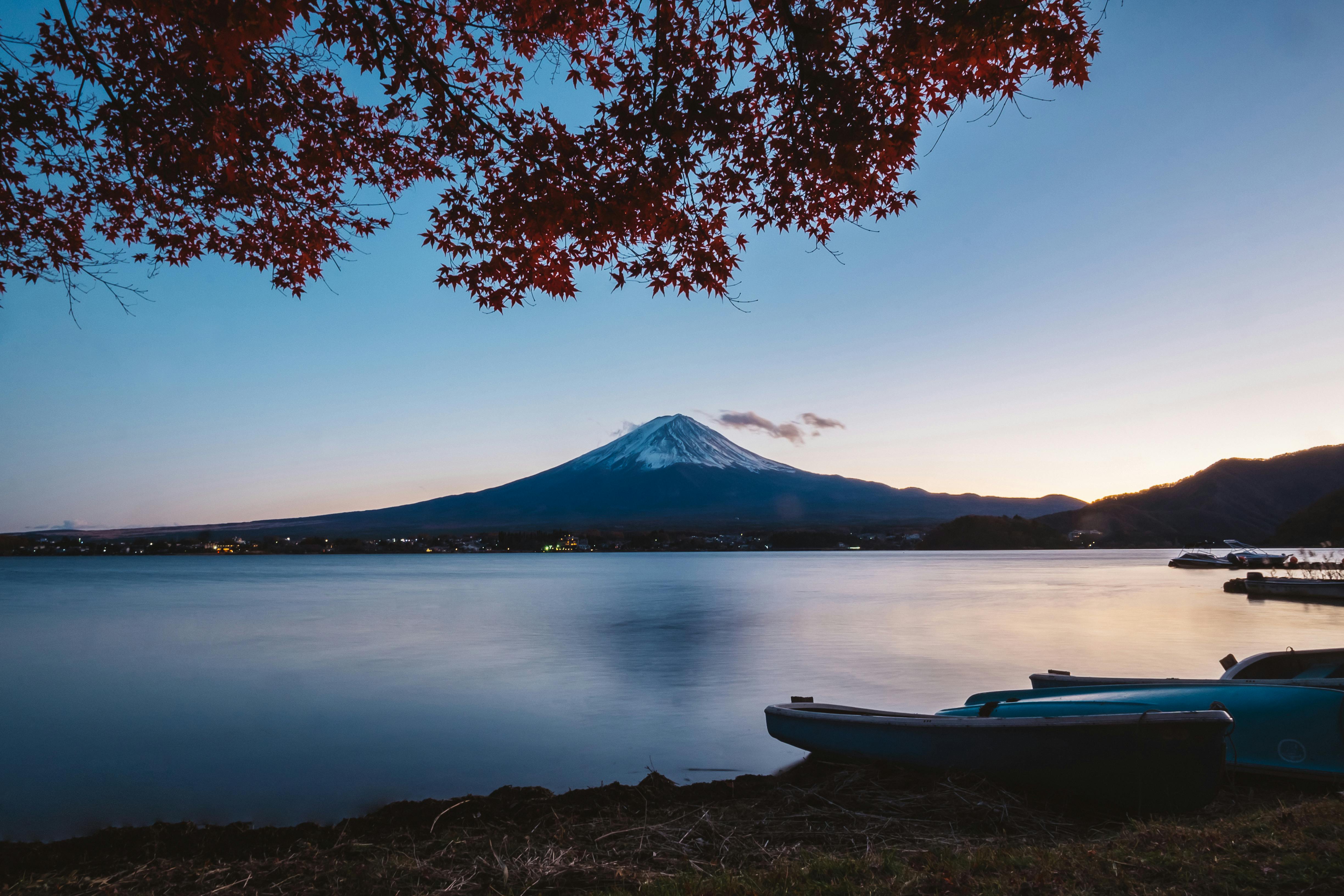 Mount Fuji at dusk, framed by vibrant red autumn leaves with small boats docked on the calm water in the foreground.