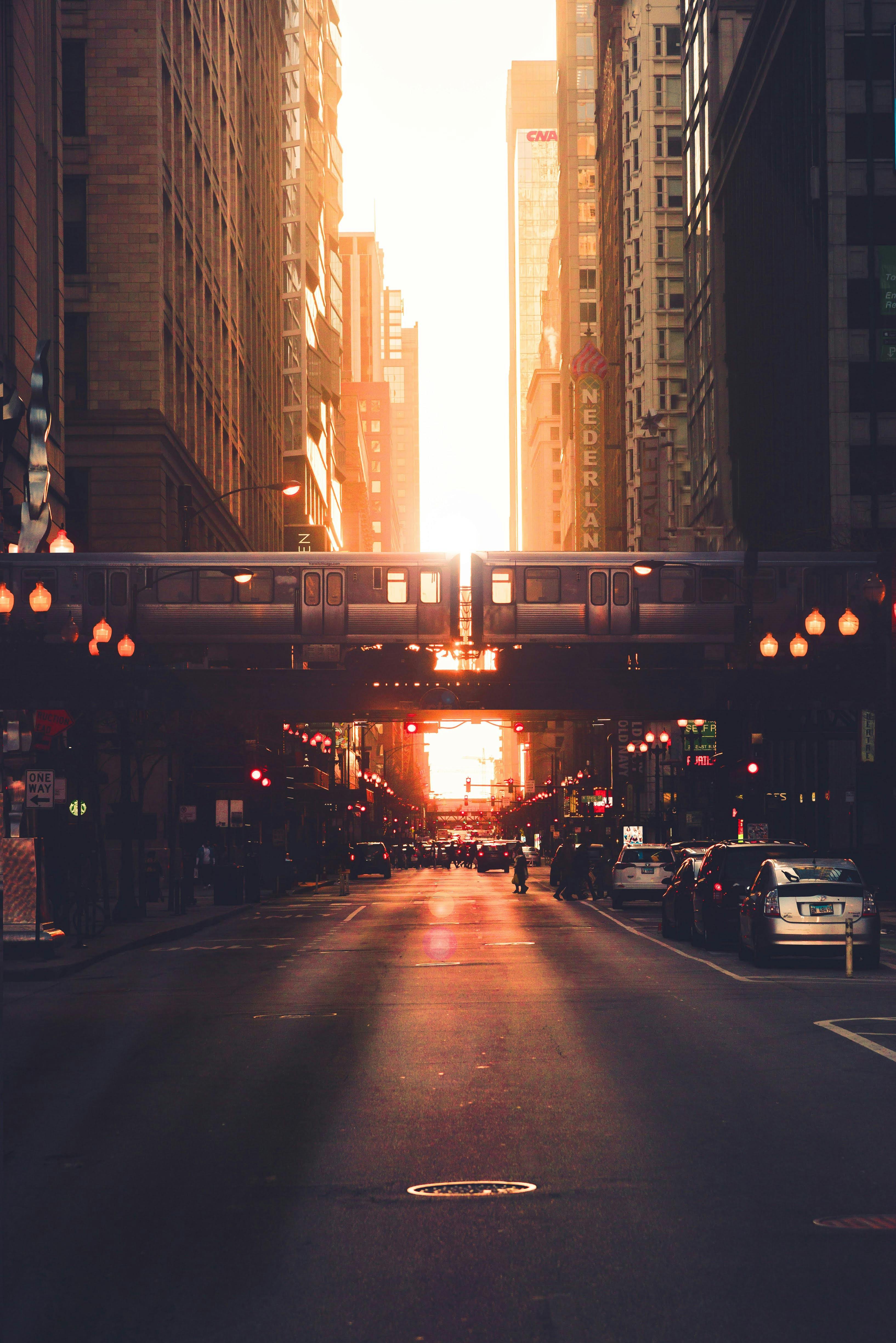 An elevated train passing over a city street at sunset, with golden light bursting through the buildings and illuminating the traffic below.