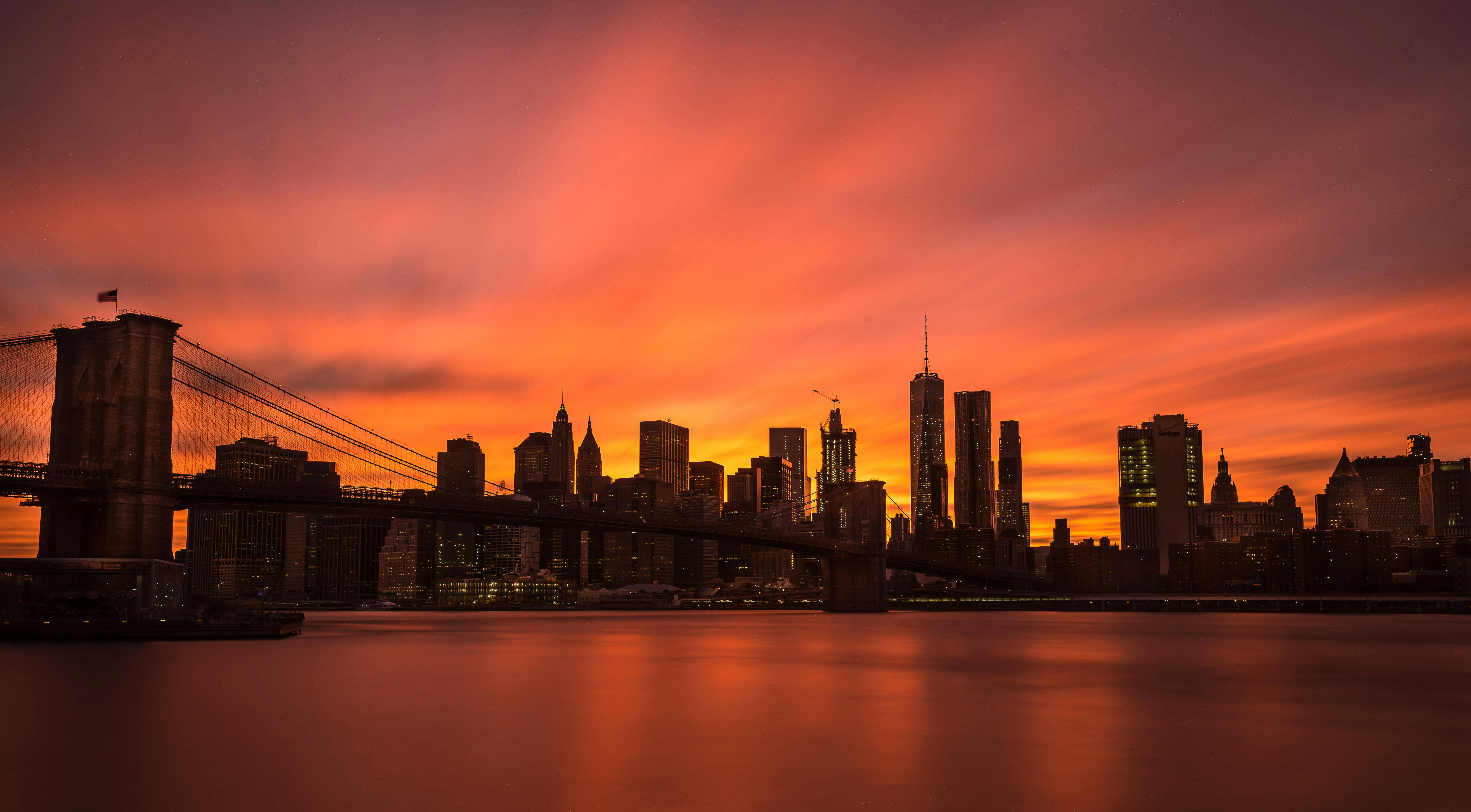 A panoramic view of the New York City skyline and Brooklyn Bridge silhouetted against a fiery orange and yellow sunset.